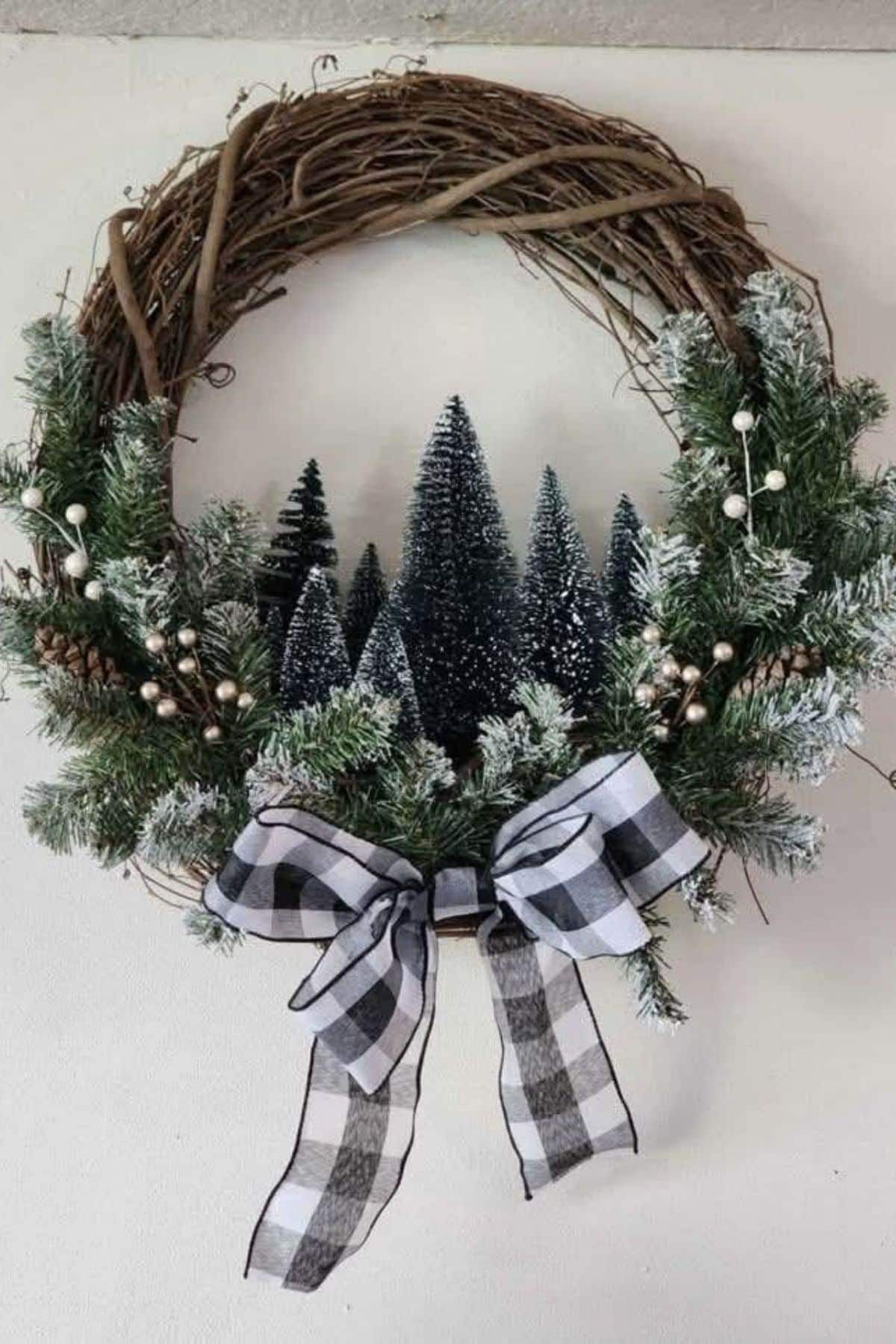 A grapevine wreath with frosted pine branches, small artificial pine trees, white berries, and a black-and-white plaid bow at the bottom, hung on a white wall.