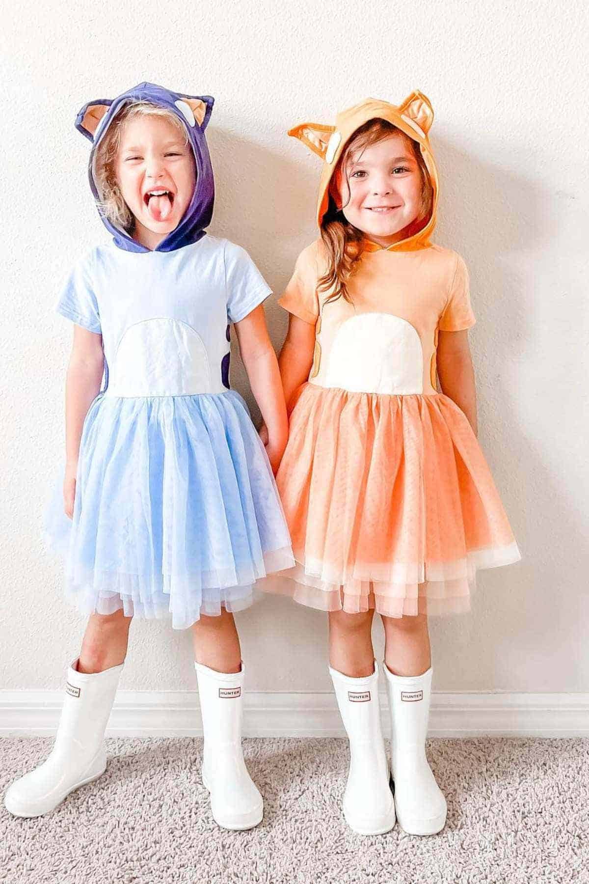 Two young children in animal-eared hooded dresses, one blue and one orange, standing side by side and wearing white rain boots against a plain wall.