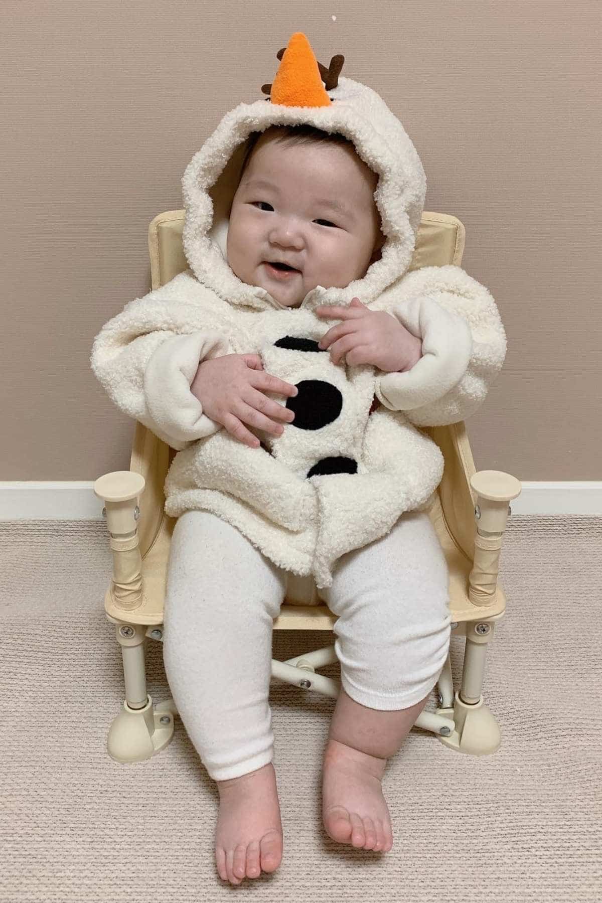 A baby sits on a small chair wearing a hooded outfit that resembles a snowman, featuring an orange “carrot” nose and black buttons.