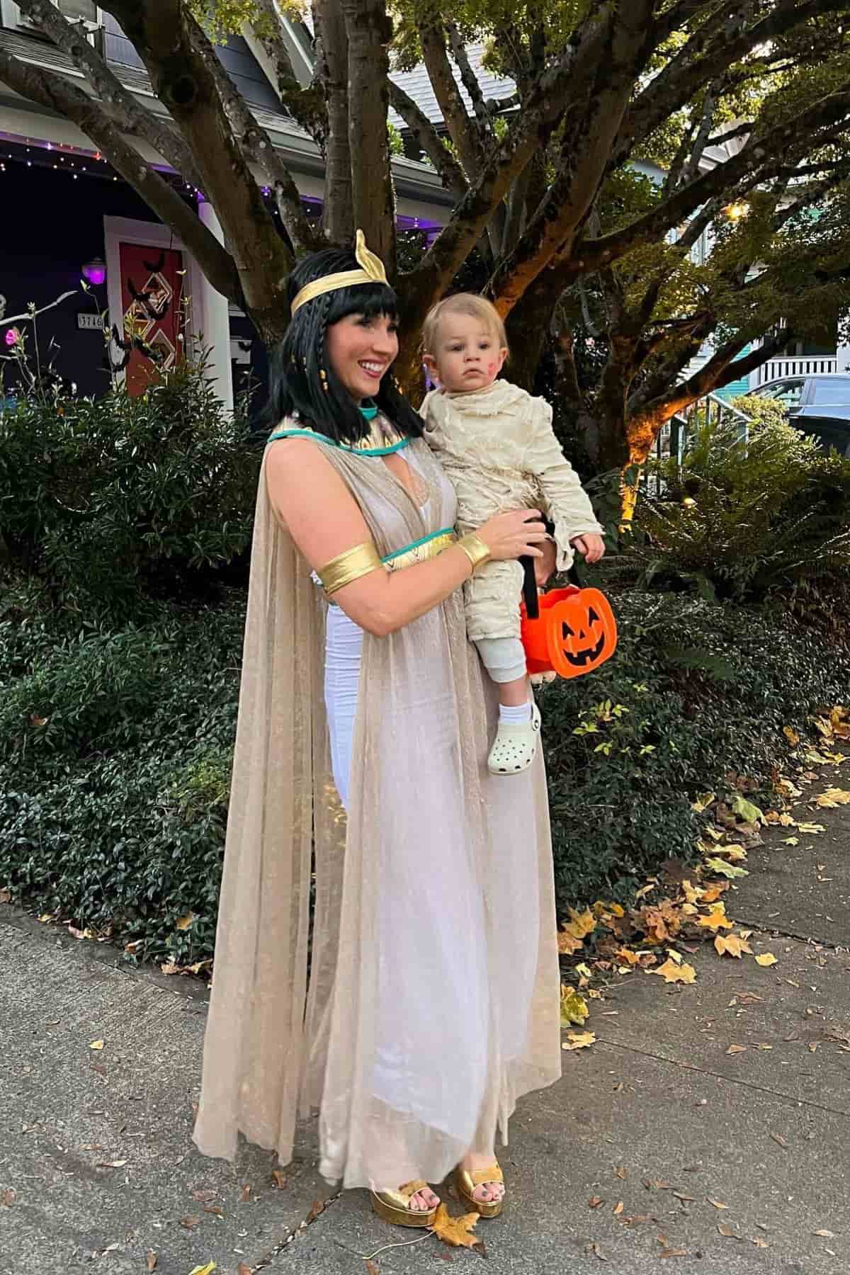 Woman in a Cleopatra costume holds a toddler dressed as a mummy with a pumpkin bucket, standing outside on a leaf-strewn sidewalk near bushes and a tree.
