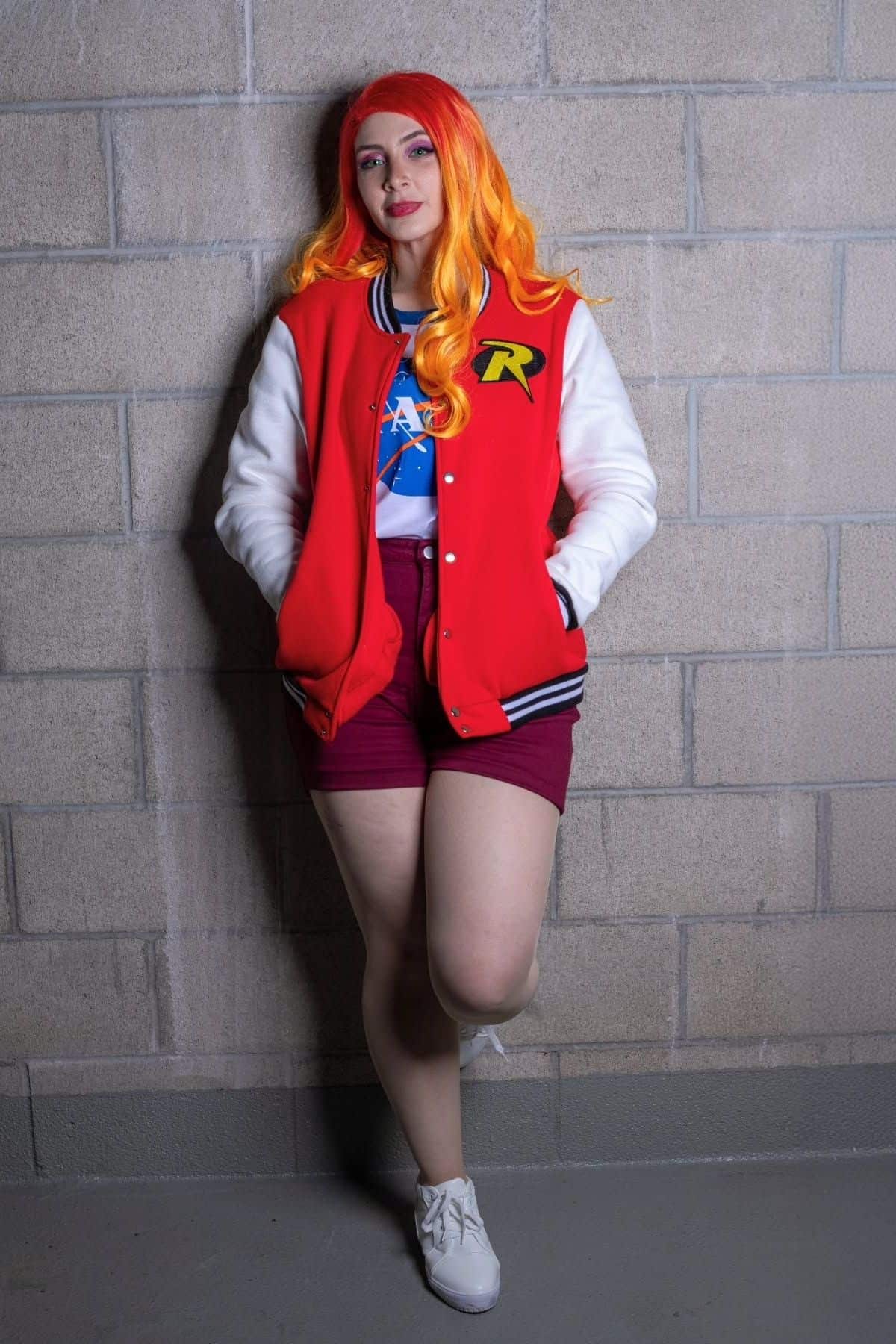 Person with orange and yellow hair, red and white varsity jacket, NASA t-shirt, red shorts, and white shoes stands against a gray brick wall with hands in pockets.