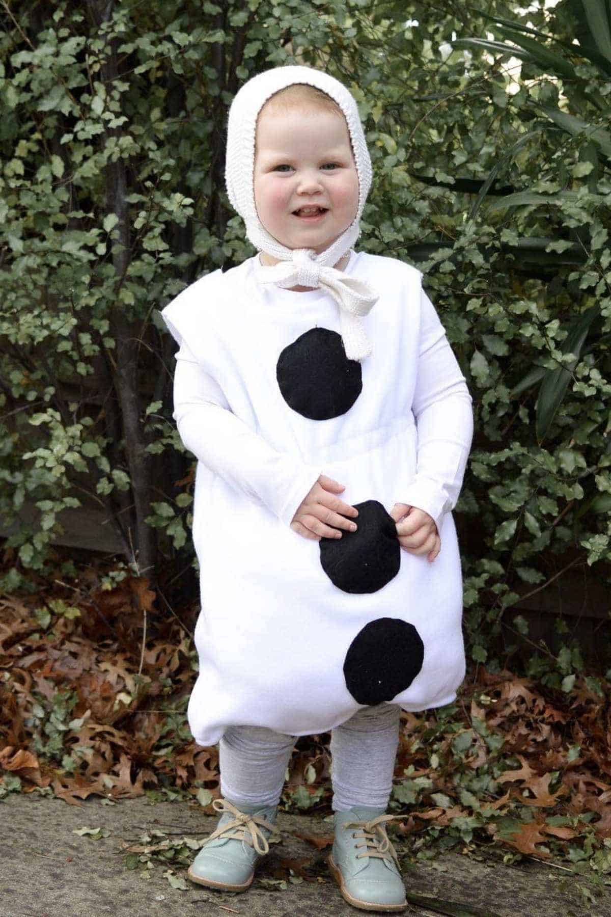 A young child stands outside wearing a white costume with three large black circles, a knit bonnet, gray leggings, and blue shoes, surrounded by greenery and fallen leaves.