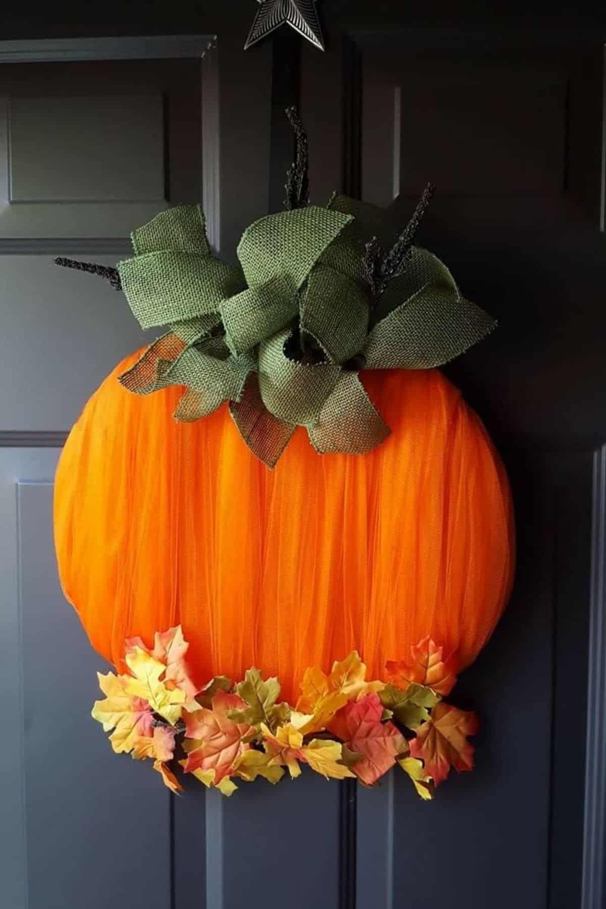 Large orange pumpkin-shaped wreath with green ribbon leaves and artificial autumn leaves at the bottom, hanging on a dark door.
