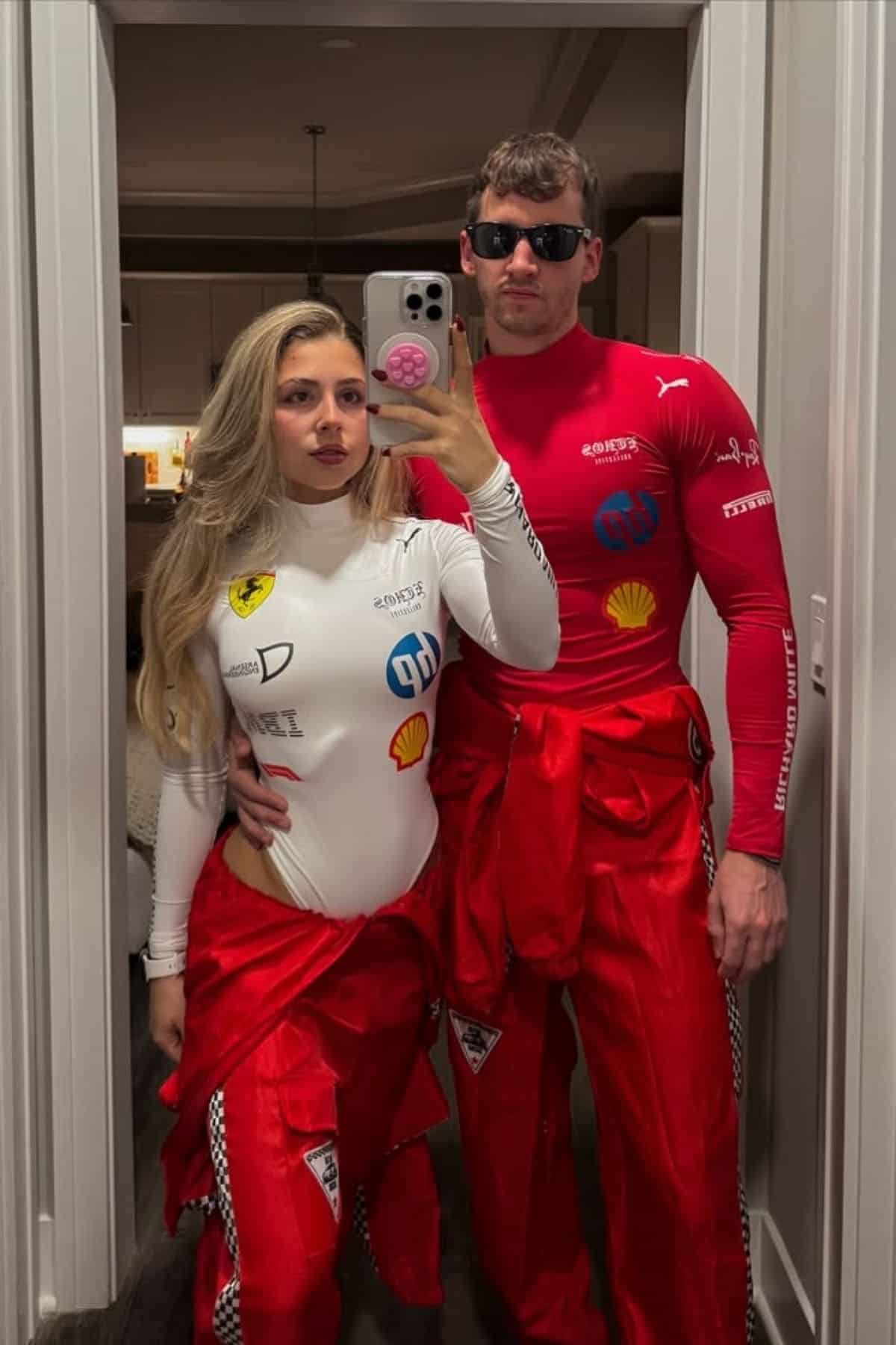 Two people in racing-themed outfits stand together in front of a mirror, posing for a photo. The person on the left wears white, and the person on the right wears red with sunglasses.