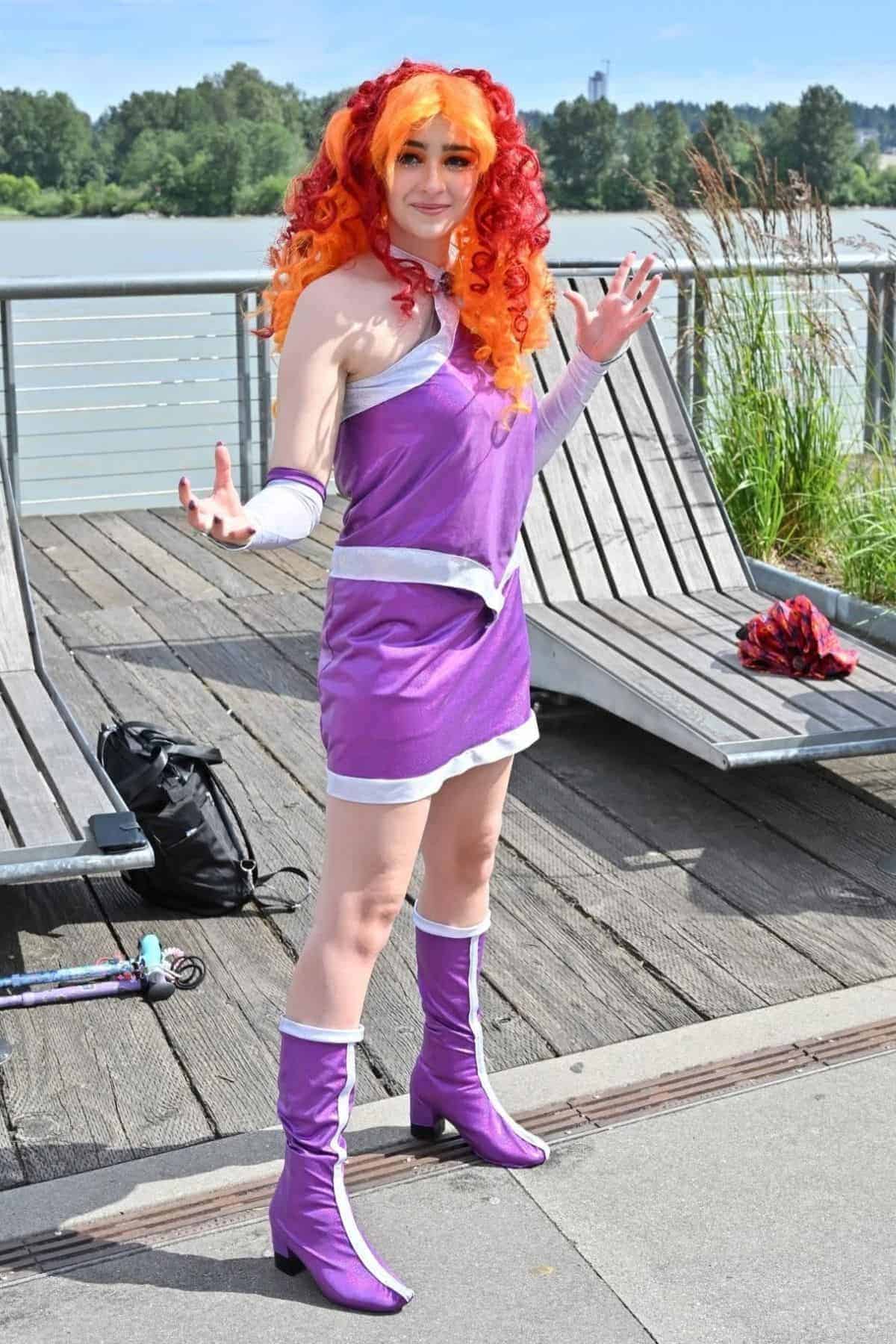 Person in a purple and silver cosplay outfit with orange and red curly hair poses outdoors near benches and a river.