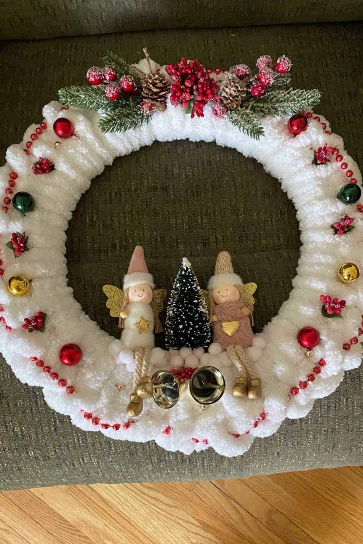 A white holiday wreath decorated with red beads, ornaments, pinecones, faux berries, two small angel dolls, a miniature tree, and two silver bells, displayed on a green surface.