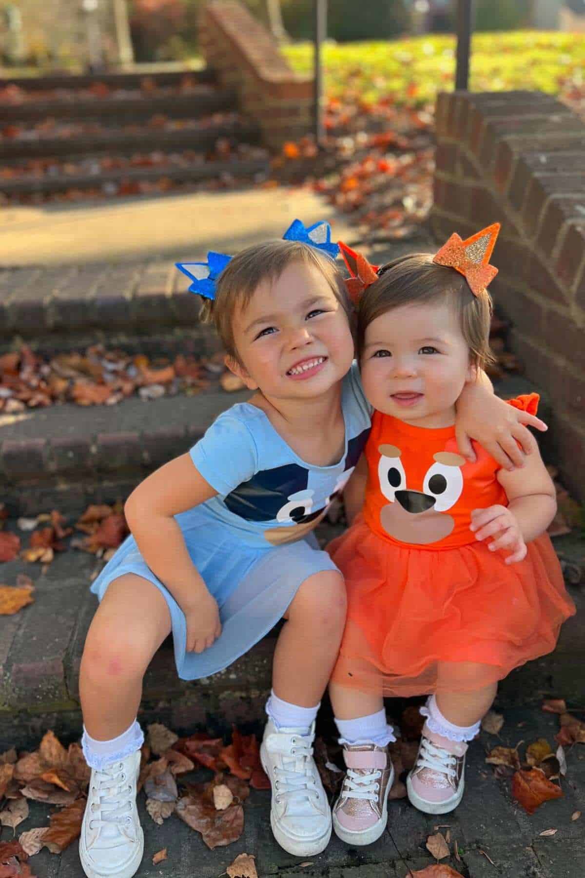 Two young children in colorful animal-themed dresses sit on outdoor steps, smiling and hugging, surrounded by autumn leaves.