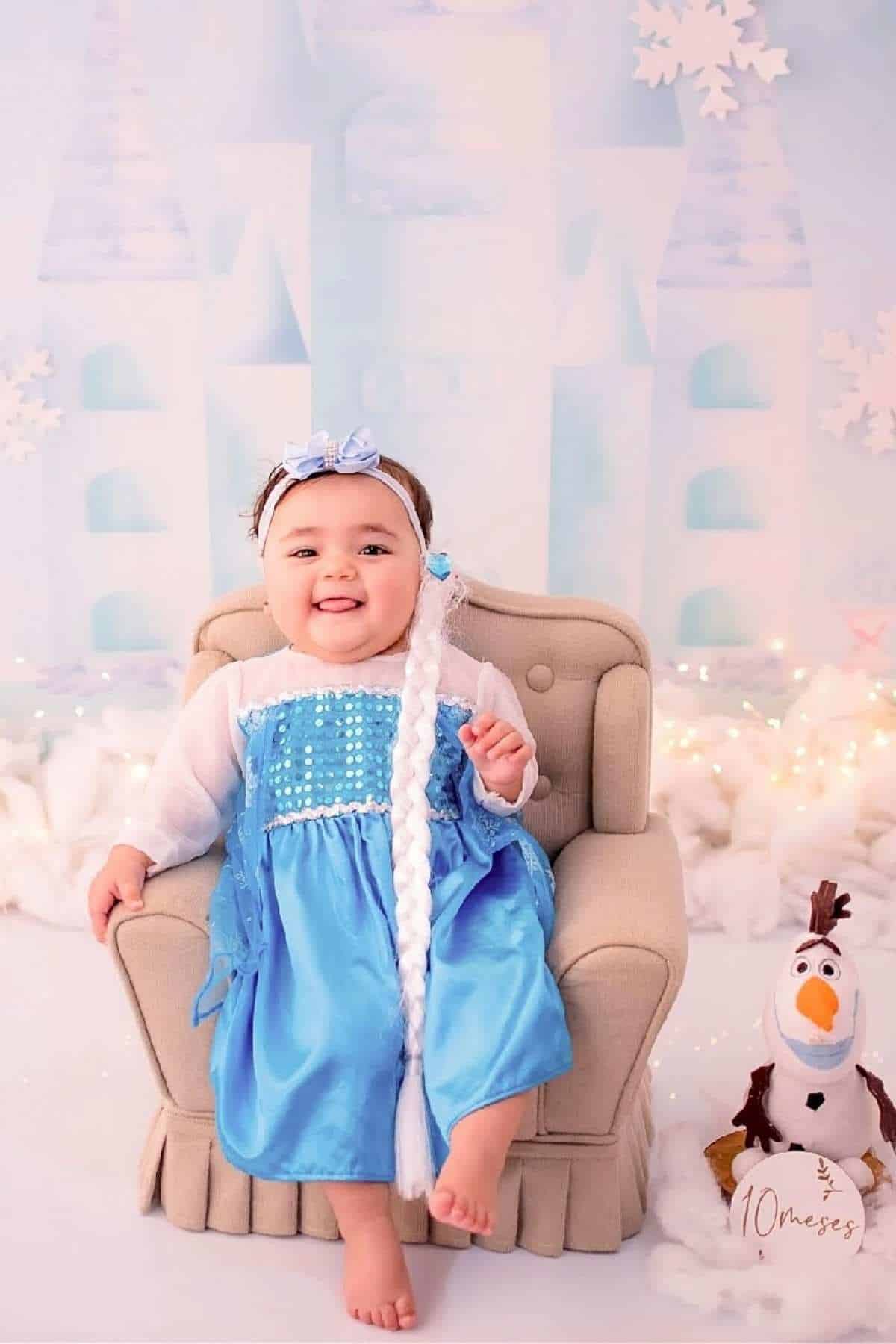 A smiling baby in a blue princess dress sits on a beige chair with a plush snowman toy beside them, against a winter-themed backdrop with snowflakes and soft decorations.