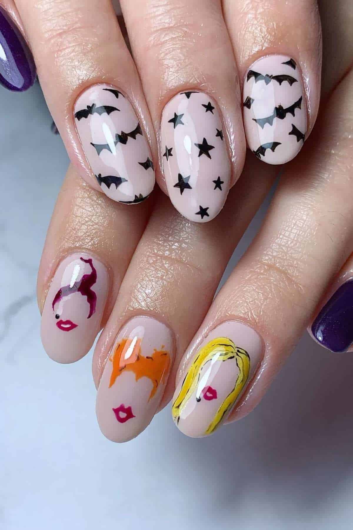 A hand with manicured nails features bats, stars, and stylized faces with red, orange, and yellow hair drawn on a light pink base.