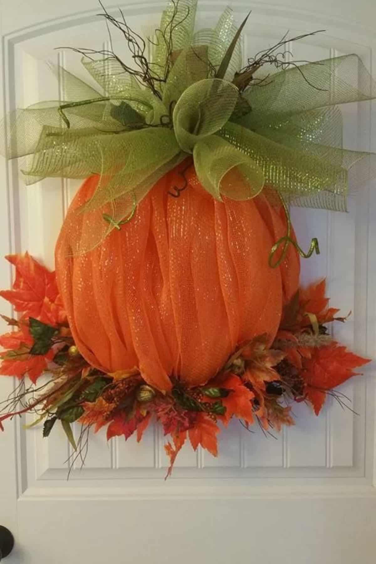 A pumpkin-shaped wreath made of orange mesh with a green mesh bow and artificial autumn leaves hangs on a white door.