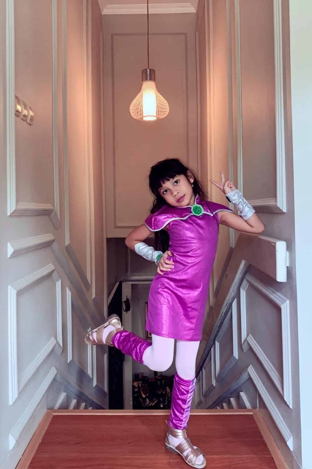 A young girl in a shiny purple costume poses playfully on a landing, with one leg raised and one hand pointing up, inside a well-lit hallway.