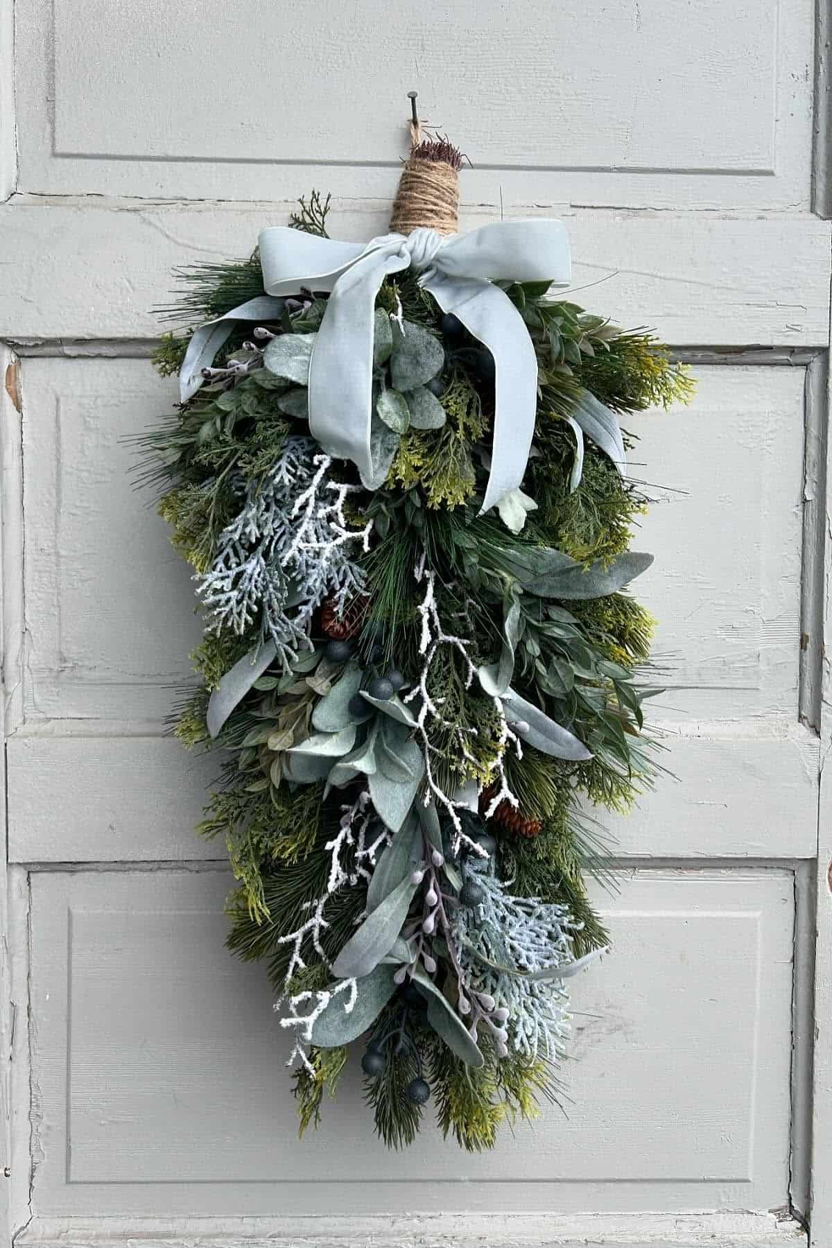 A swag of mixed greenery, frosted branches, and pinecones, tied at the top with twine and a white ribbon, hangs on a light gray wooden door.