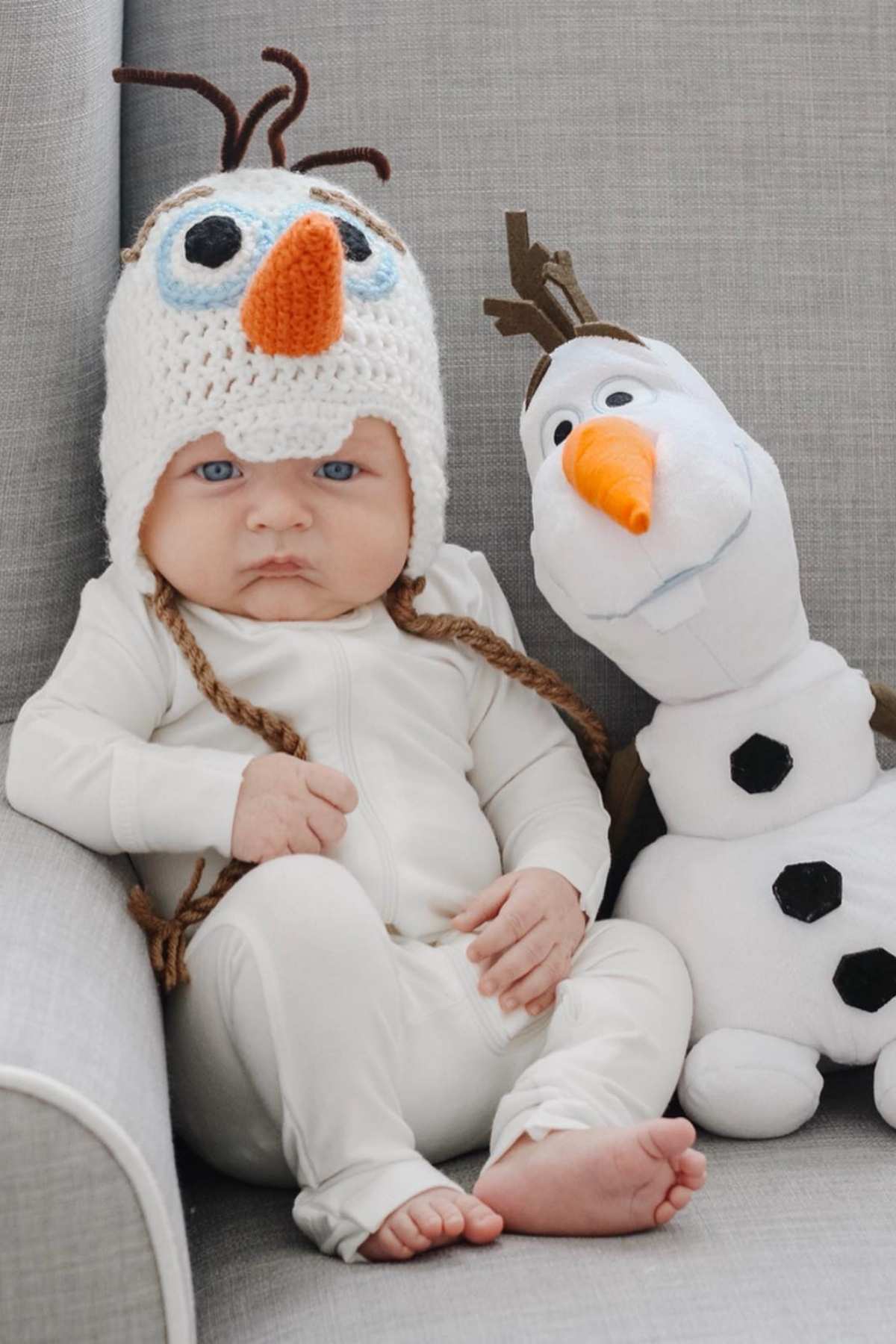 Baby in a white outfit wearing a crocheted snowman hat sits on a gray chair next to a plush snowman toy.