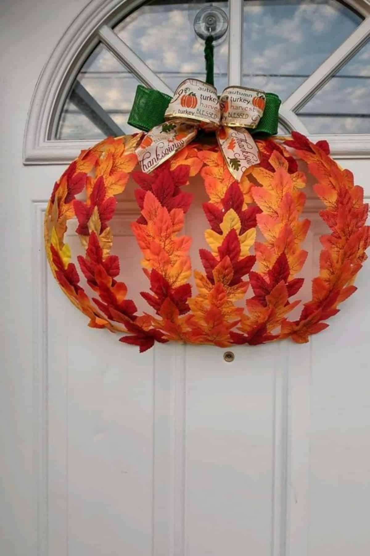 A pumpkin-shaped wreath made of orange, red, and yellow leaves with a decorative ribbon bow hangs on a white door.