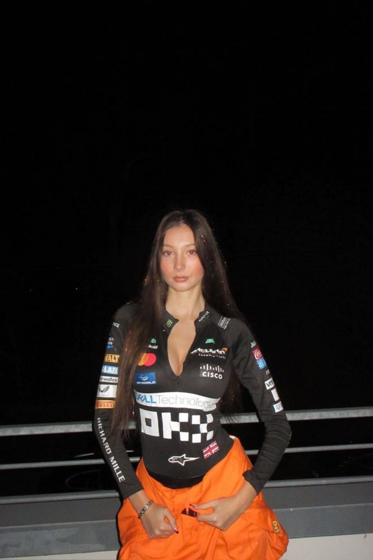 A woman with long brown hair wears a black racing jacket with sponsor logos and orange pants, standing against a dark background and holding the waistband of her pants.