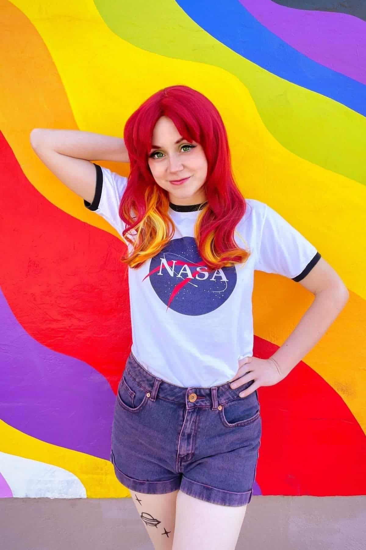 Person with red and yellow hair poses in front of a colorful, wavy rainbow mural, wearing a NASA t-shirt and denim shorts with hand on hip.