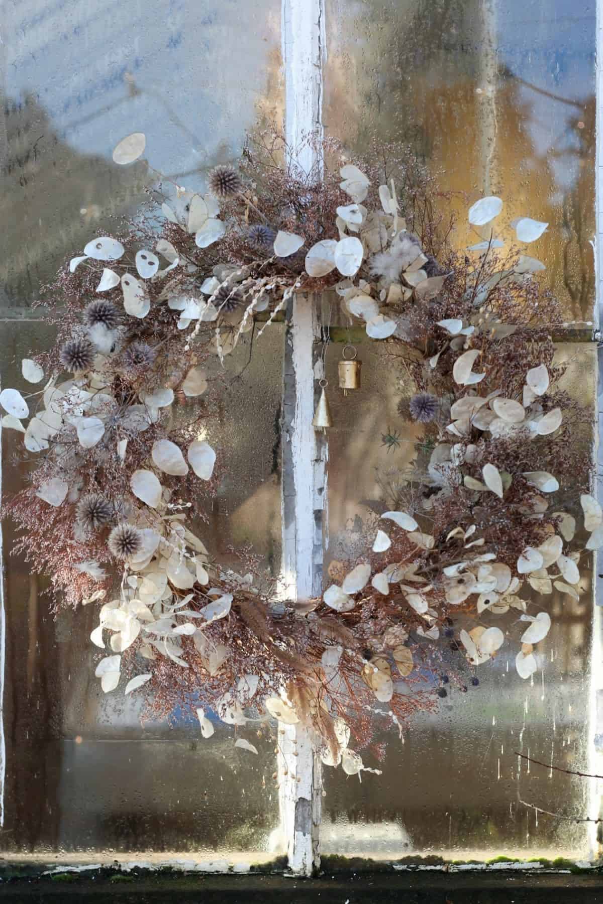 A wreath made of dried white and brown foliage hangs on a weathered, foggy window with peeling paint.