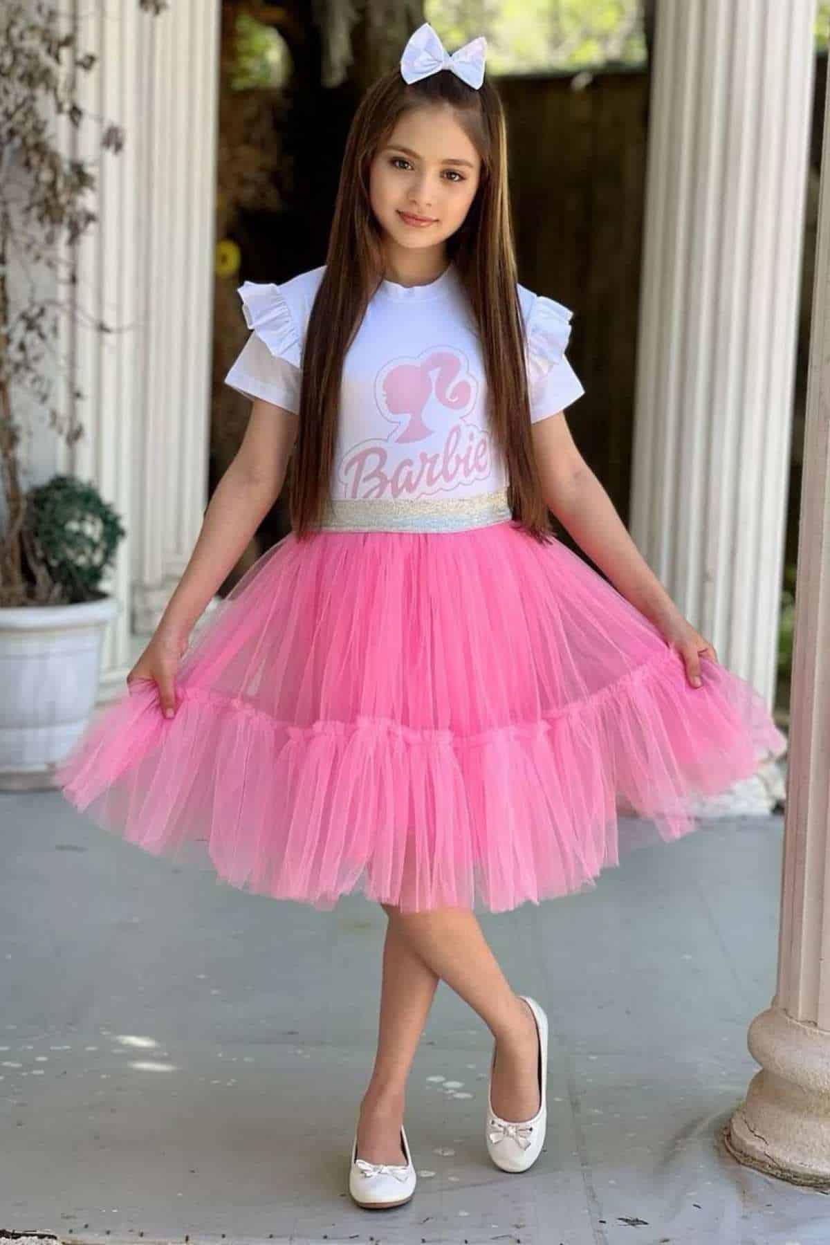 Young girl stands on a porch wearing a white shirt with “Barbie” print, a pink tulle skirt, white bow headband, and white shoes. She poses with one foot crossed in front of the other.
