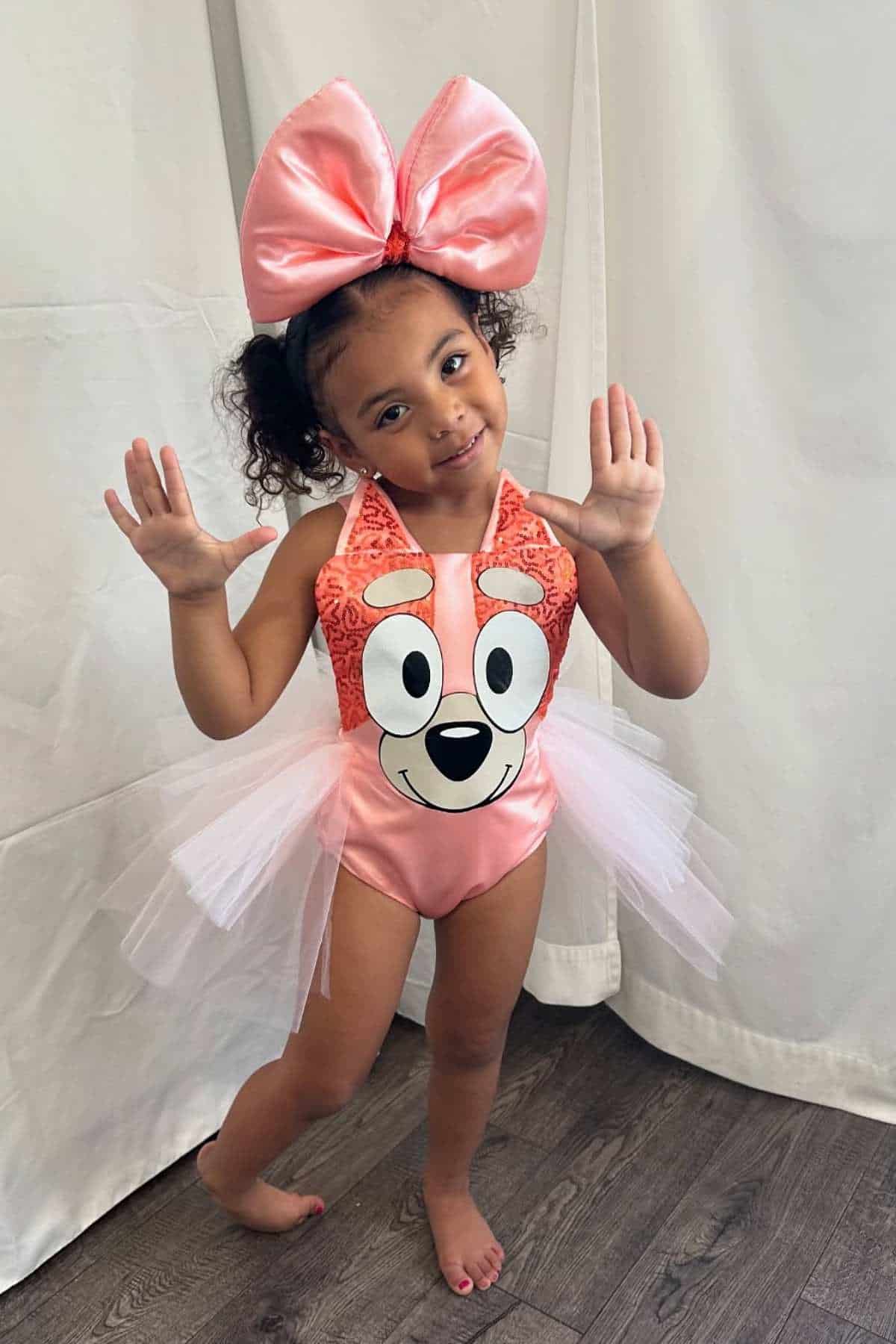 Young child poses indoors wearing a pink cartoon-themed dress with a large matching bow and tulle skirt, standing on wooden flooring in front of a light curtain.