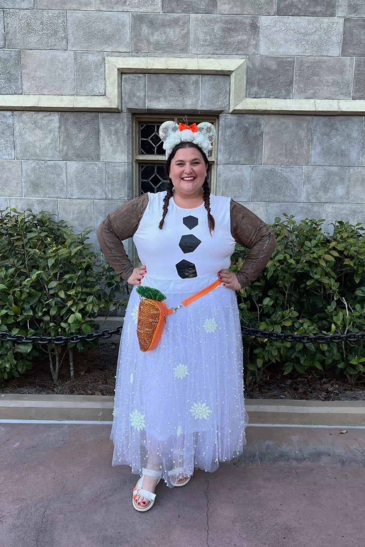 Person in a snowman-inspired costume with brown sleeves, white dress with snowflakes, carrot-shaped purse, and mouse-ear headband with an orange bow, standing outside against a stone wall.