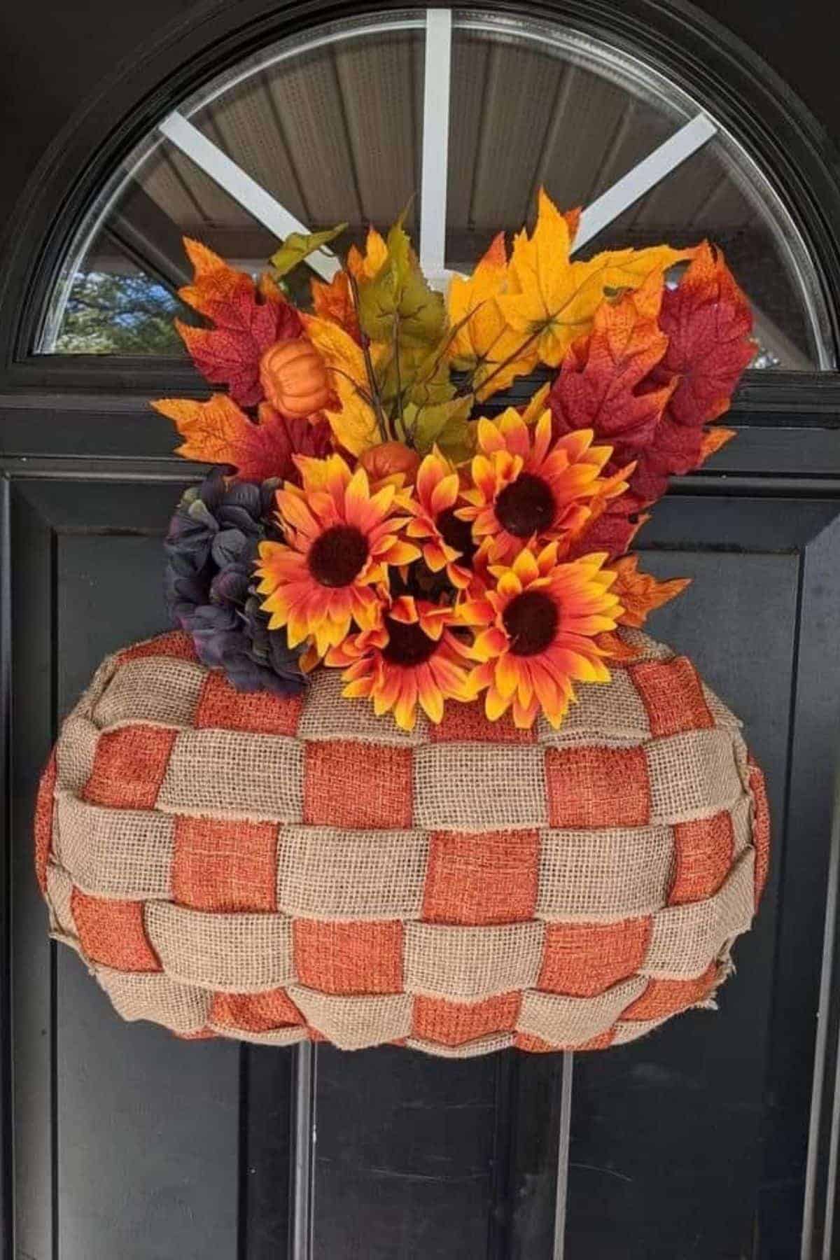 Woven basket of burlap and orange fabric holding artificial sunflowers, pumpkins, and fall leaves hangs on a black front door.
