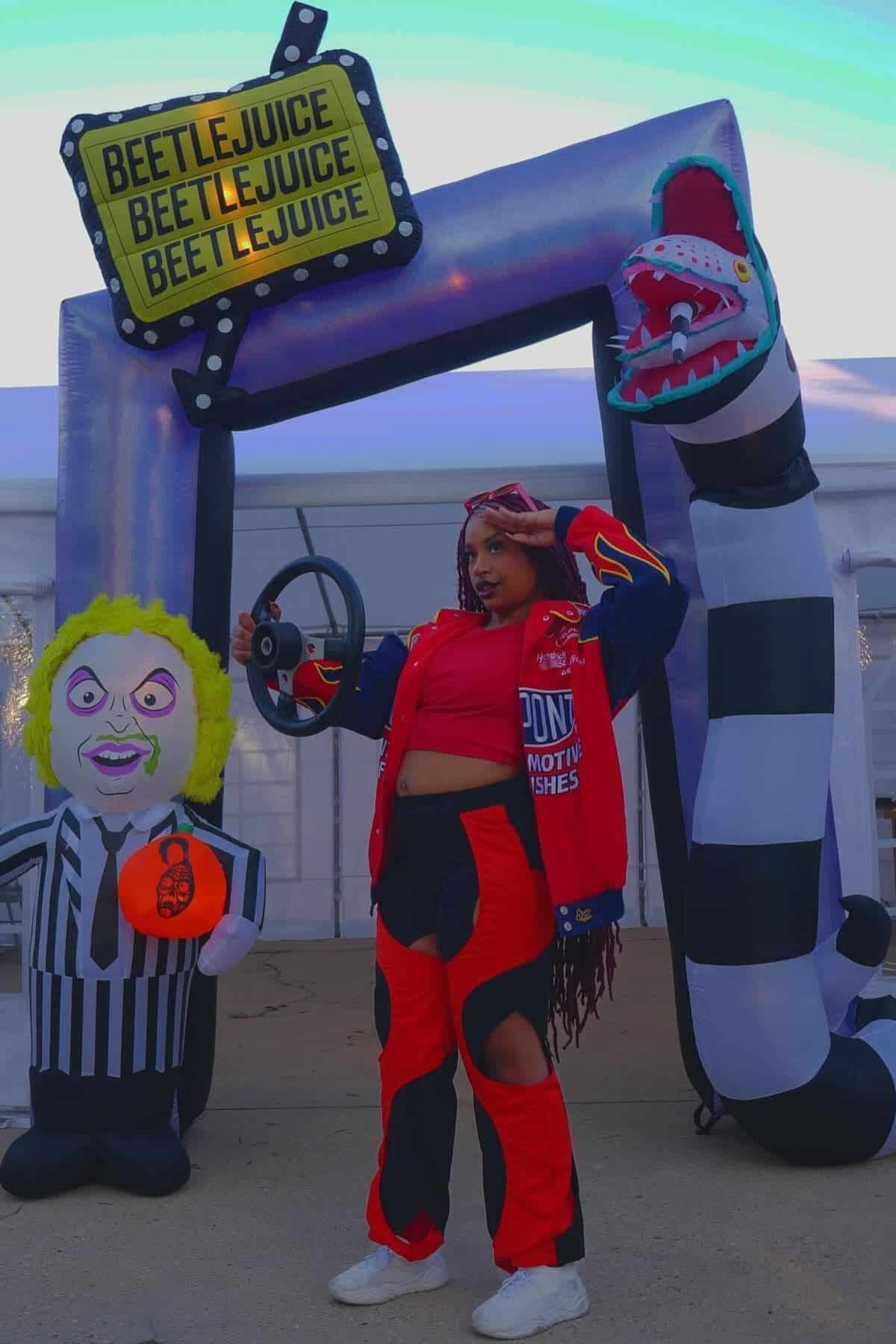 Person in a red and black racing outfit poses in front of Beetlejuice-themed inflatable decorations, including a sign, a striped character figure, and a sandworm.