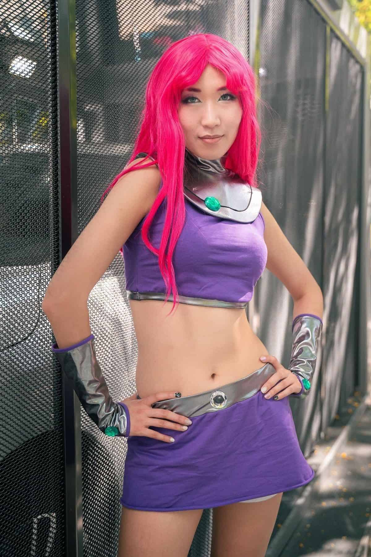 Person in a purple costume with pink hair and metallic accessories stands outdoors in front of a wire fence, posing for the camera.