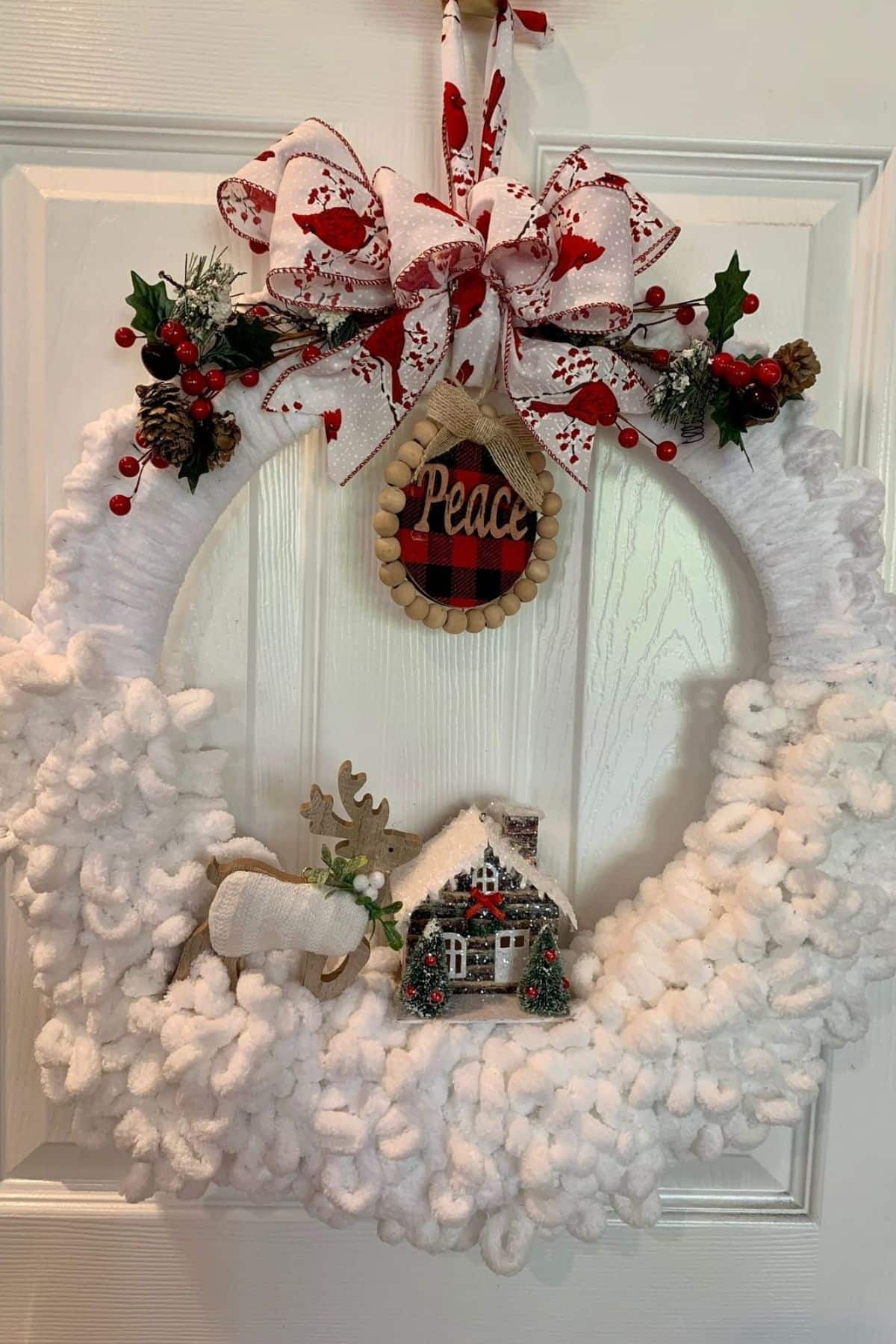 A white, fluffy Christmas wreath with red berries, greenery, a plaid “Peace” sign, a reindeer, and a small house, hanging on a white door.