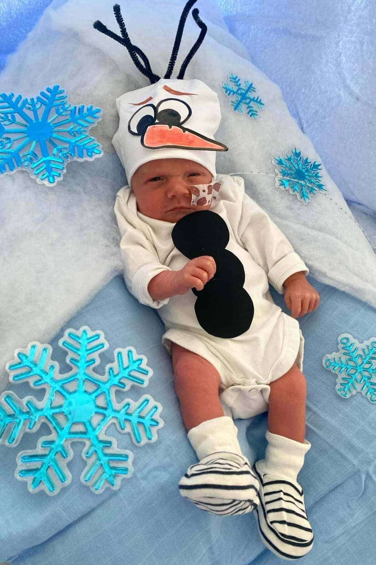 Newborn dressed in a snowman-themed outfit with black buttons, striped booties, and a hat featuring a snowman face, lying on a blue and white blanket with snowflake decorations.