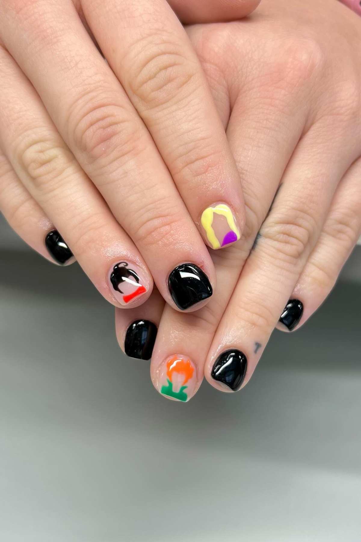 Hands with short, black-painted nails featuring colorful abstract designs on the thumbs and pinky nails, resting on a light surface.