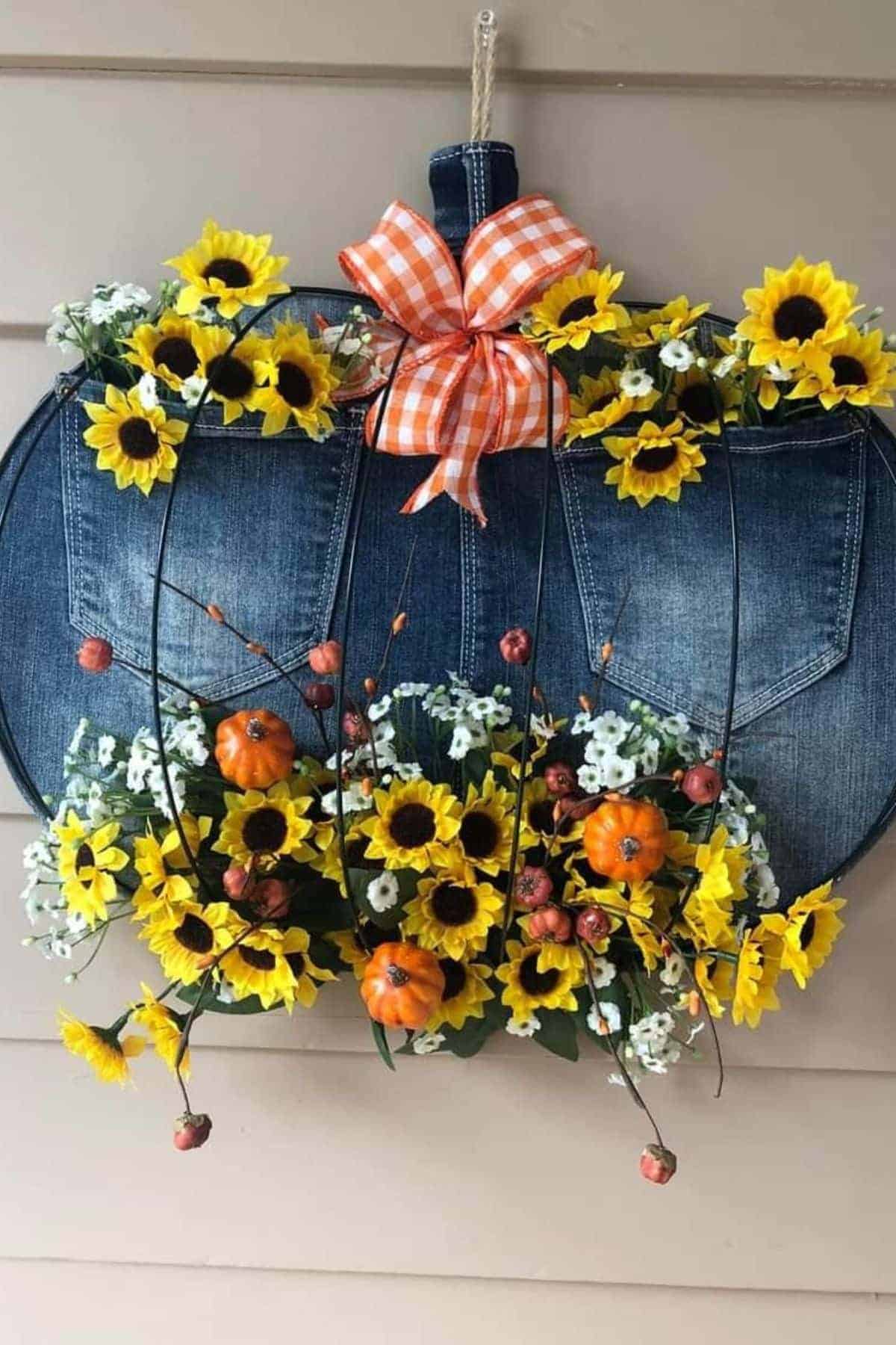 A pumpkin-shaped wreath made from denim with sunflower, pumpkin, and berry decorations, topped with an orange gingham bow, hangs on a beige wall.