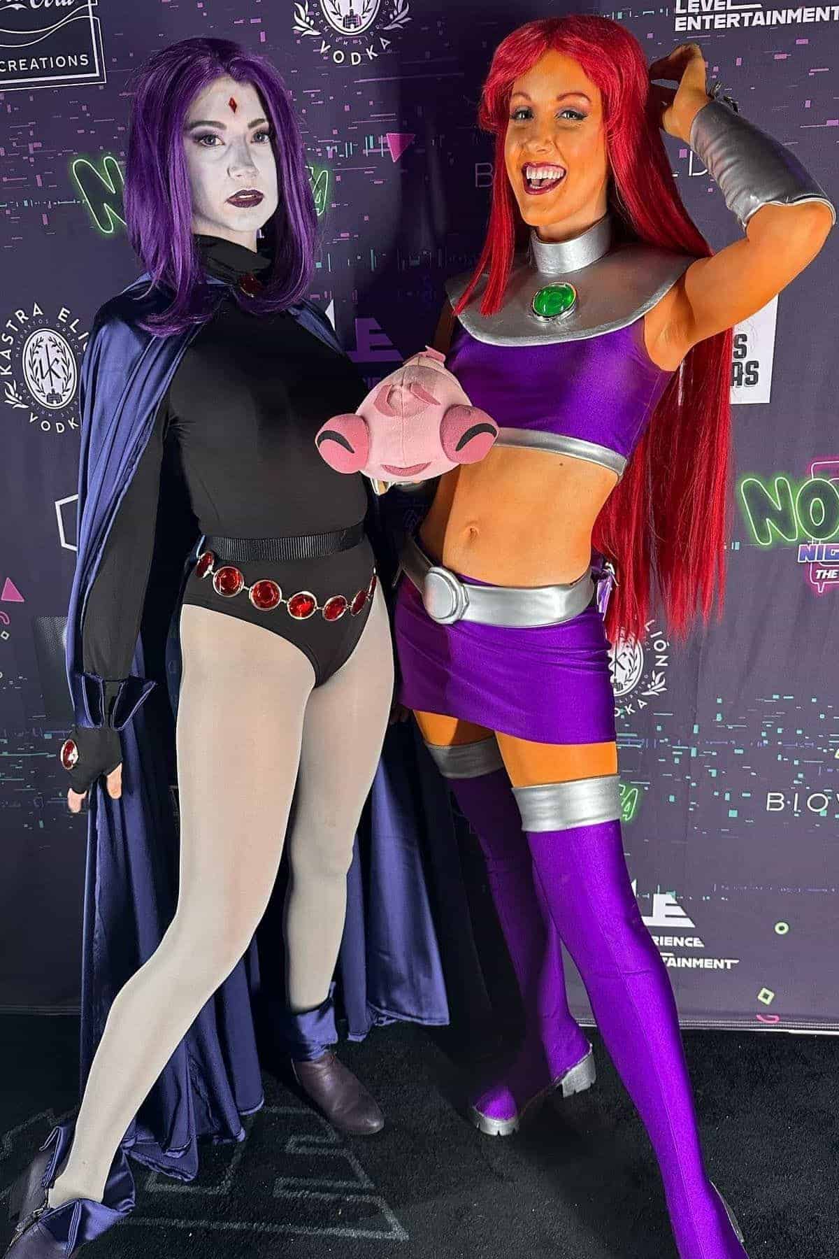 Two women in detailed Raven and Starfire costumes pose together at an event with a themed step-and-repeat backdrop.