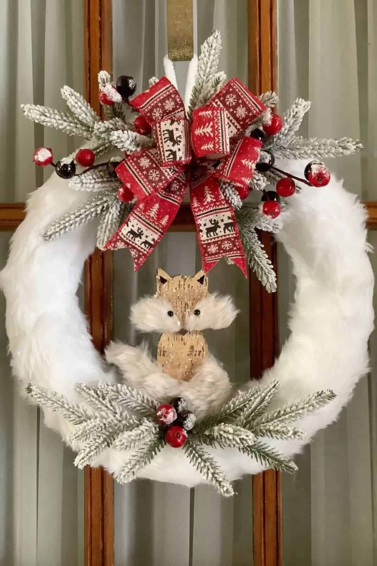A white holiday wreath with frosted pine branches, red berries, a red patterned bow, and a small fox figure in the center, hanging on a wooden door.