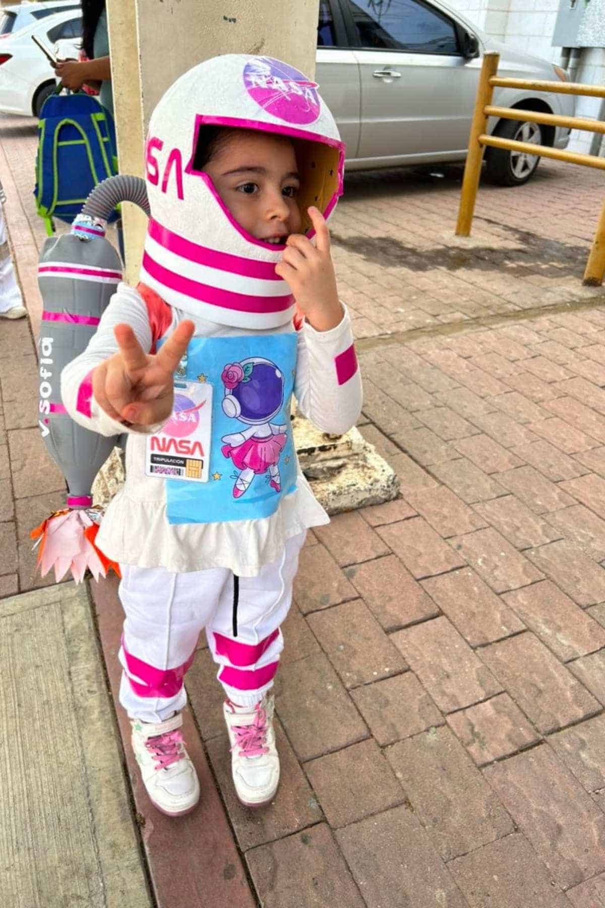 A young child in a homemade astronaut costume with pink accents stands on a sidewalk, holding up two fingers in a peace sign.