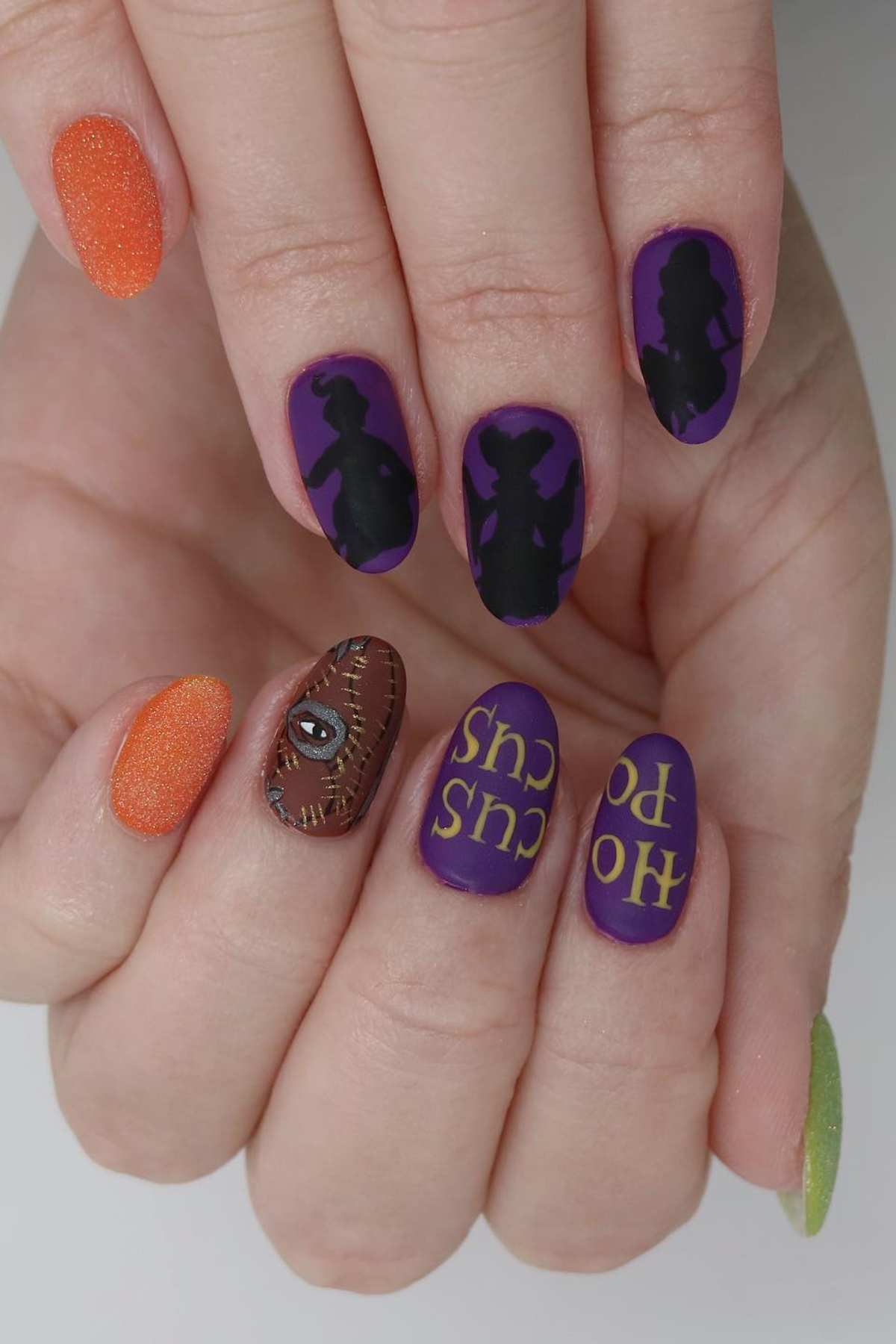 A hand with colorful Halloween-themed nail art featuring witch silhouettes, the phrase “Hocus Pocus,” an eyeball, and orange and green glitter polish.