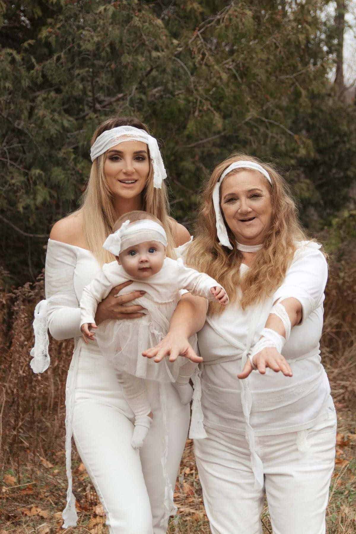Three people dressed in white mummy costumes pose outdoors; an adult woman holds a baby, and another woman stands beside them, both with arms outstretched toward the camera.