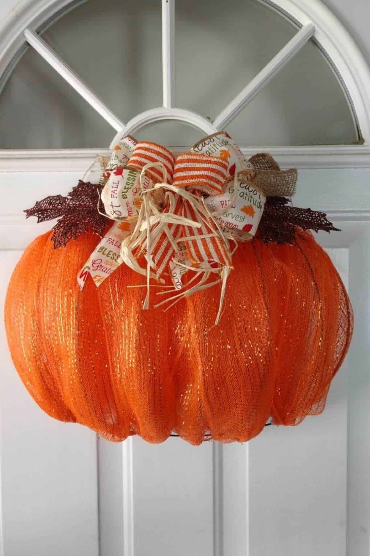 Orange mesh wreath shaped like a pumpkin hangs on a white door, decorated with fall-themed ribbons, raffia, and faux leaves.