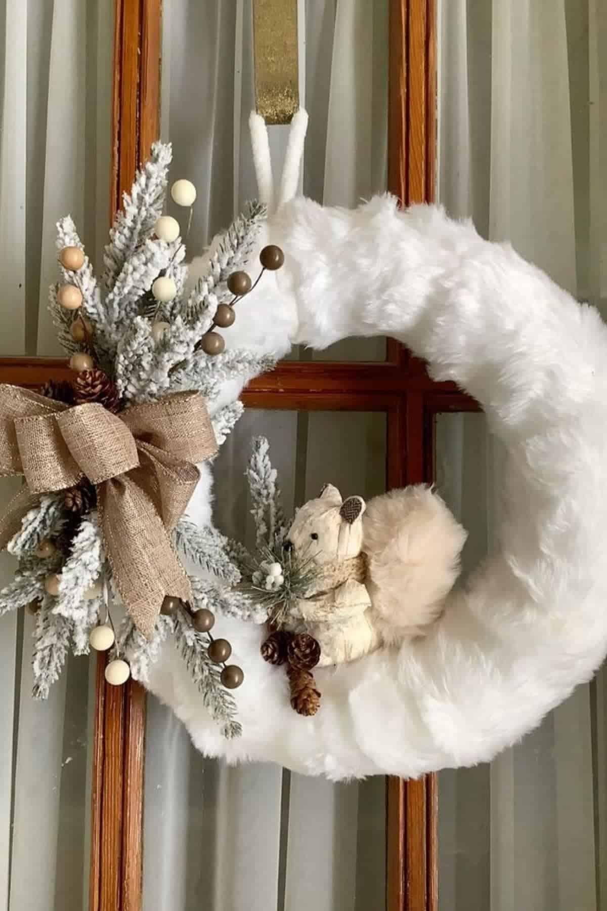 A white faux fur wreath decorated with a burlap bow, frosted greenery, berries, pinecones, and a small squirrel figurine hangs on a glass-paned door.