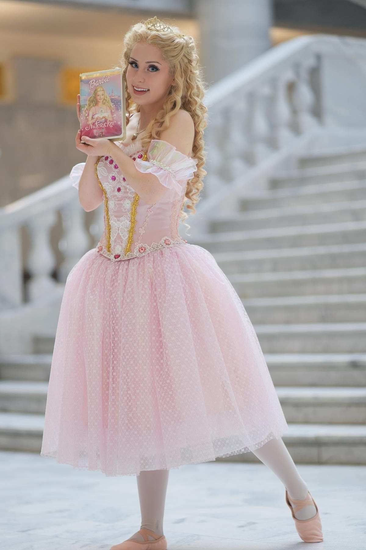 Woman in a pink princess costume poses on stairs, holding up a Barbie Nutcracker DVD with both hands and smiling at the camera.