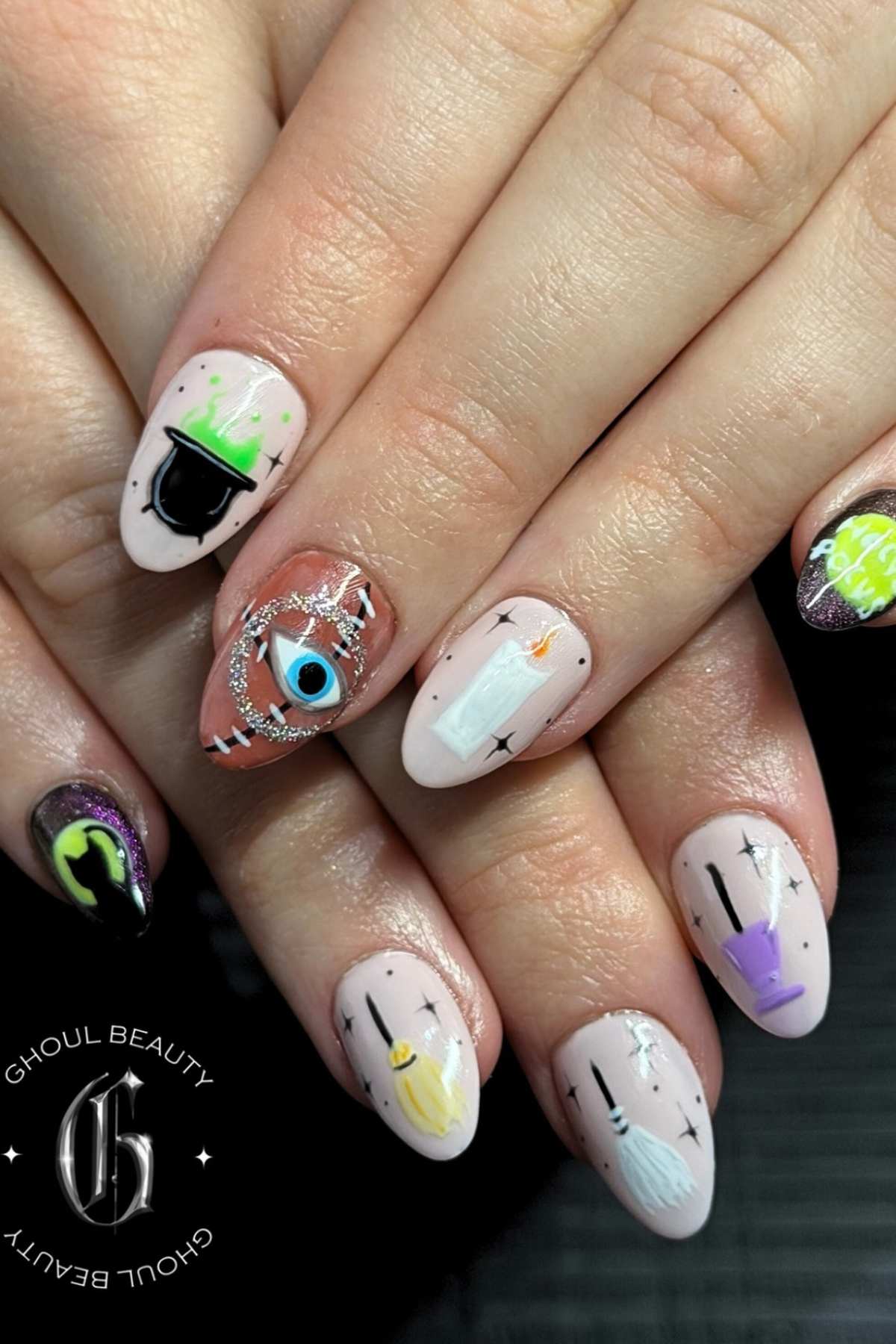 Close-up of hands with Halloween-themed nail art, including candles, a cauldron, an eye, potion bottles, and sparkles on almond-shaped nails. Ghoul Beauty logo is in the lower left corner.