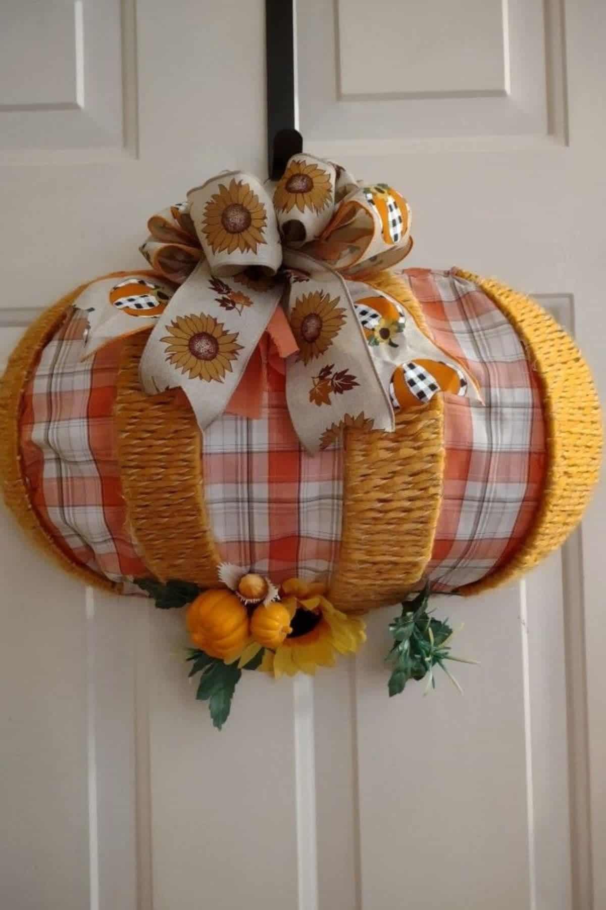 A plaid and wicker pumpkin-shaped wreath hangs on a door, decorated with a sunflower bow and small artificial pumpkins and greenery at the bottom.