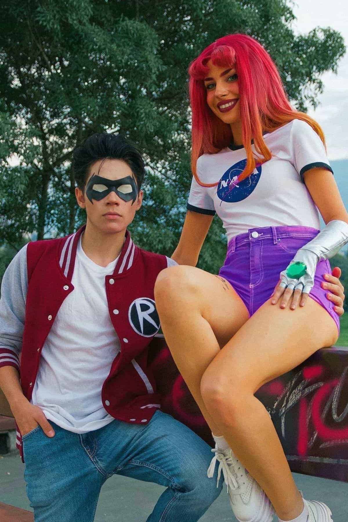 Two people in cosplay; one wears a red varsity jacket with an “R” and mask, the other has a red wig, NASA shirt, purple shorts, and a green ring. They pose outdoors in front of trees and graffiti.