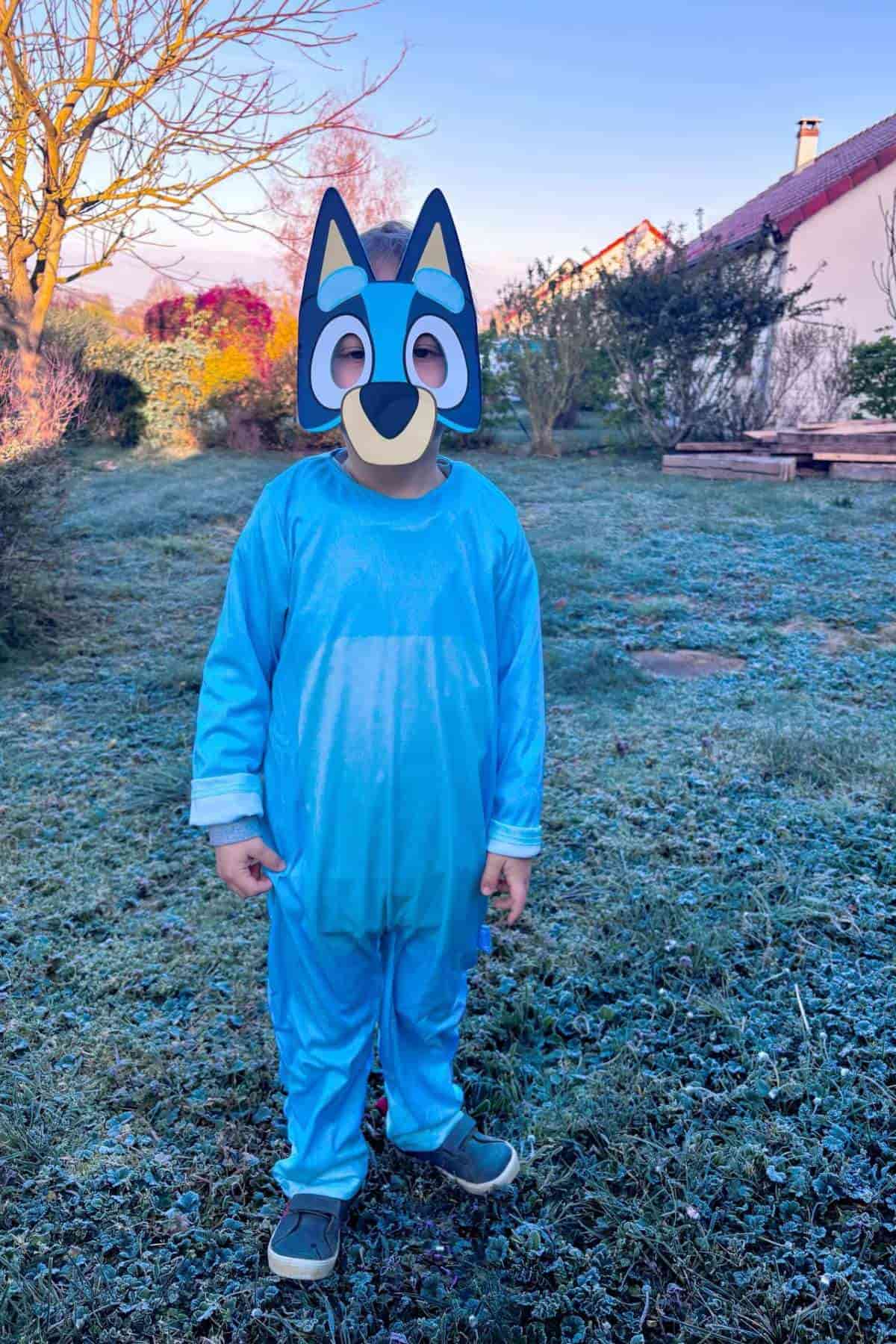 A child wearing a blue costume and a cartoon dog mask stands outside on frosty grass with houses and trees in the background.