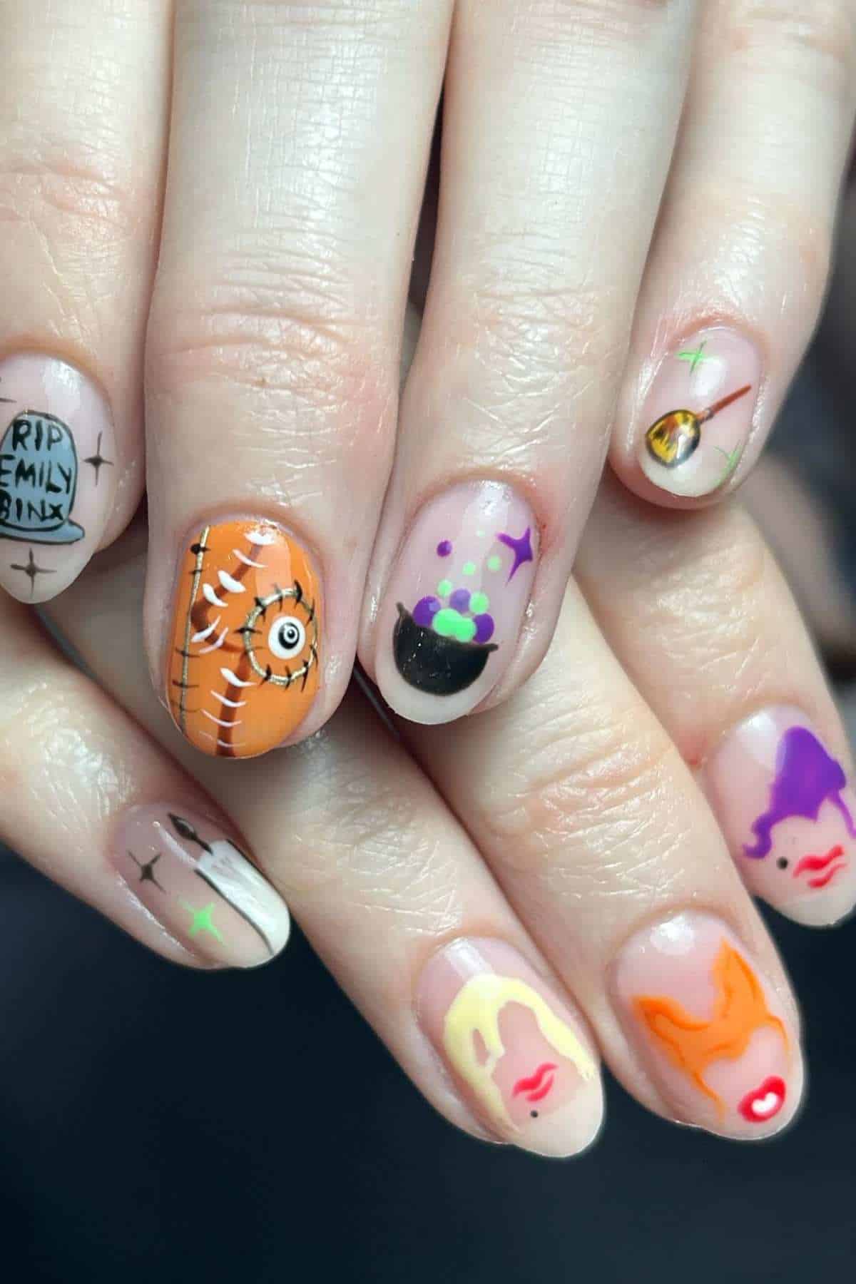A hand with Halloween-themed nail art, including a cauldron, stitched orange pattern, “RIP Emily” tombstone, gold eye, and stylized faces in bright colors on natural nails.