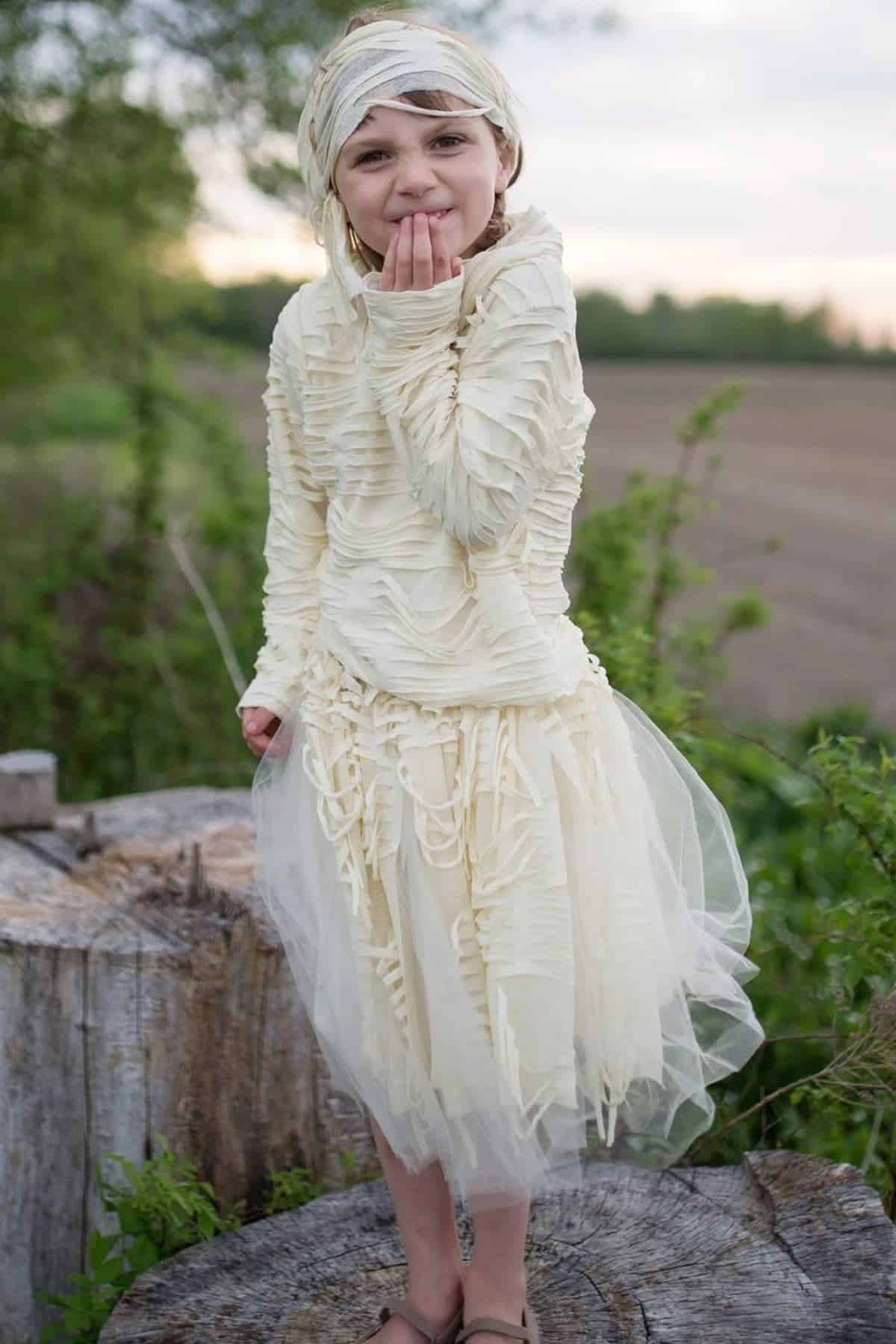 A child wearing a textured cream-colored dress and head wrap stands outdoors on a tree stump, holding one hand to their mouth.