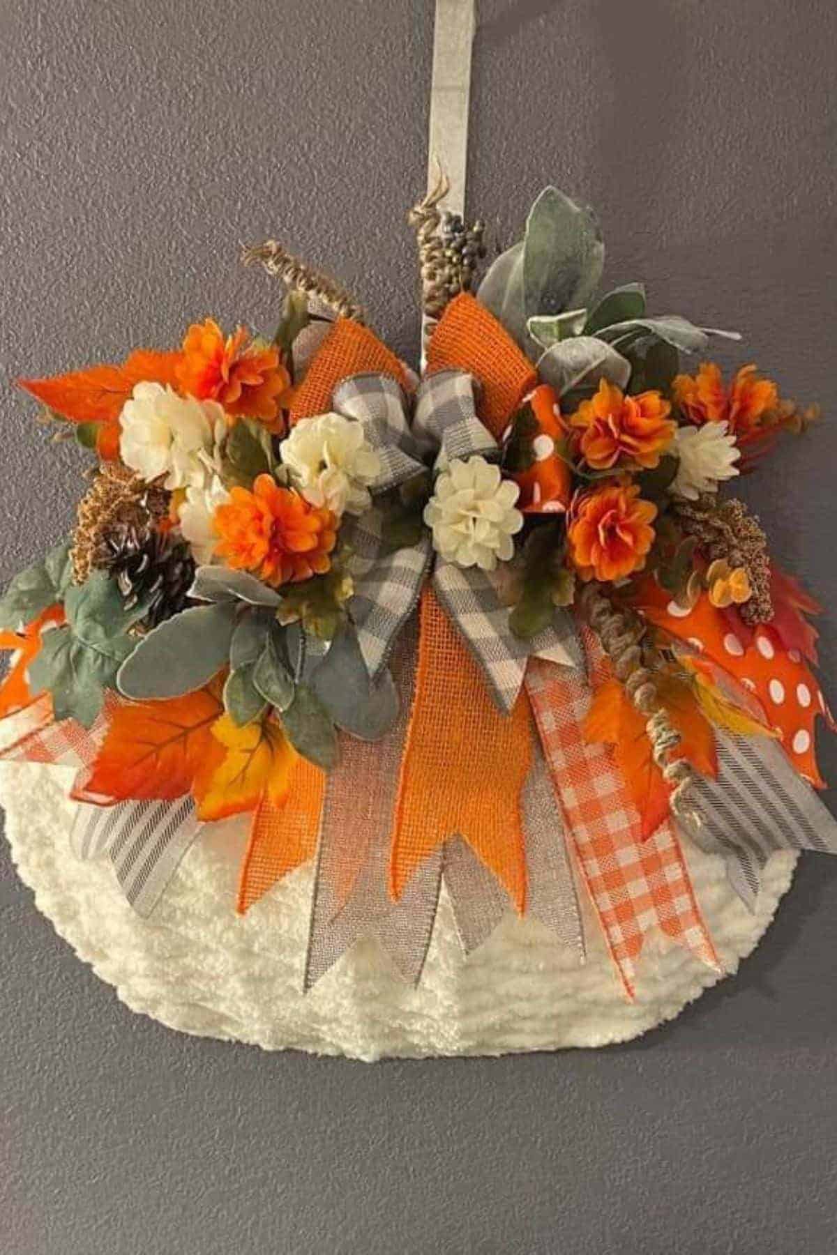 A decorative fall wreath with orange, white, and yellow flowers, assorted leaves, and plaid ribbons, hanging on a gray wall.