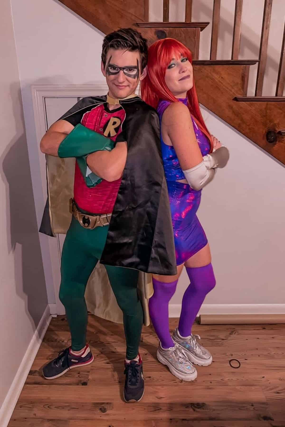 Two people in superhero costumes stand back-to-back indoors. One is dressed as Robin, the other as Starfire, both posing near a wooden staircase.
