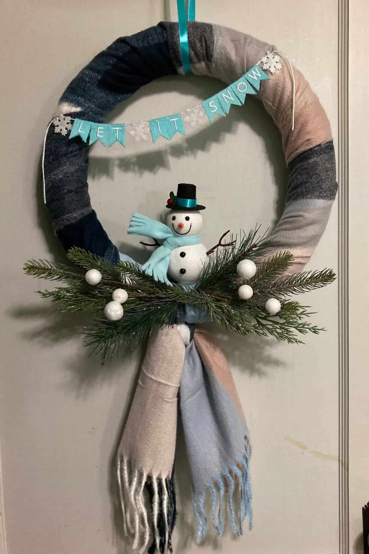 A wreath wrapped in pastel fabric features a snowman, pine branches, white berries, and a “LET IT SNOW” banner.