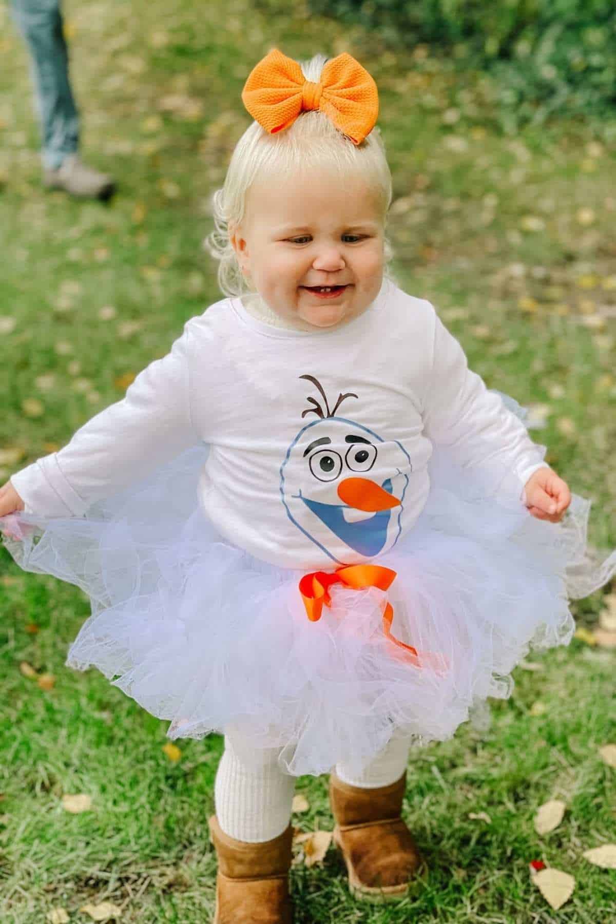 Toddler in white tutu and Olaf shirt smiling outdoors, wearing tan boots and an orange bow, standing on grass with autumn leaves.