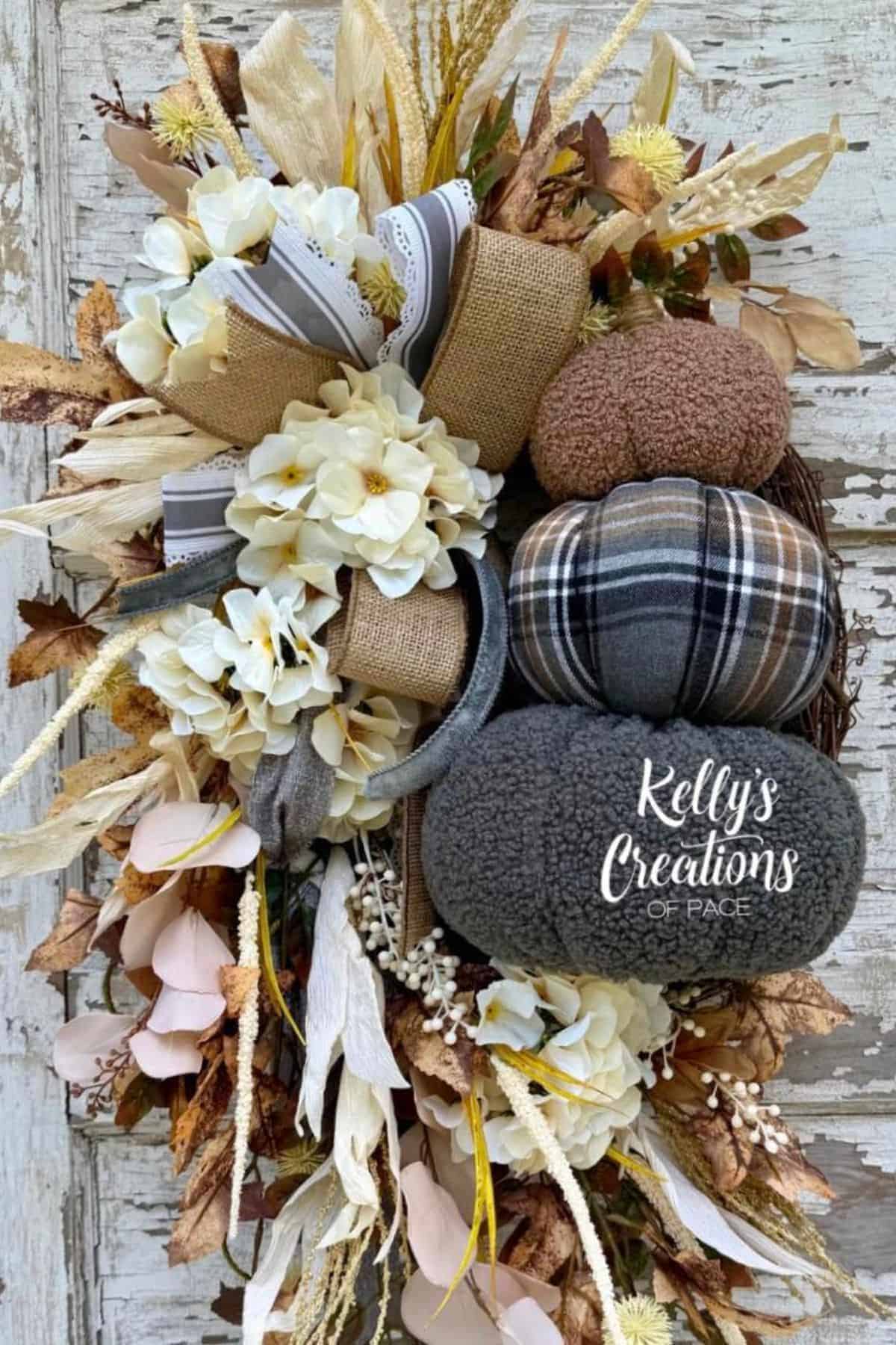 Fall wreath with fabric pumpkins, white flowers, burlap bows, and dried foliage on a rustic wooden background. “Kelly’s Creations of Pace” is written on one pumpkin.